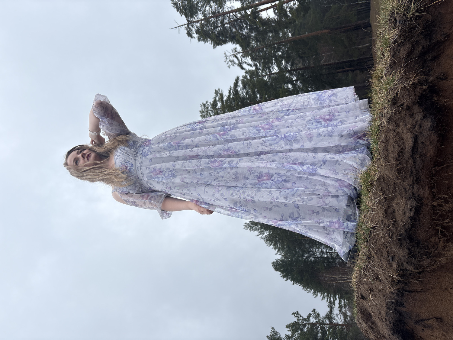 The image, currently rotated 90 degrees clockwise, depicts a woman posing outdoors. When oriented correctly, she stands on a steep dirt bank or cliff edge, looking directly towards the viewer with a neutral expression. She has long, light blonde hair and is wearing a long, flowing, pleated dress with a white or very light background adorned with a purple and blue floral pattern. The dress features sheer or lacy sleeves, and one of her arms is raised with her hand near her head, while the other is slightly bent downwards.
To the woman's left, the textured brown dirt bank rises, topped with patches of green grass. In the background, beyond the bank and the woman, a dense line of tall evergreen trees, likely pines, forms a dark, silhouetted forest edge. The upper right portion of the image is dominated by an overcast, light gray sky, indicating a cloudy daytime setting without direct sunlight. The scene suggests a natural, outdoor location, possibly at the edge of a forest or a elevated landscape in Chaikovsky, Russia. There is no visible text.