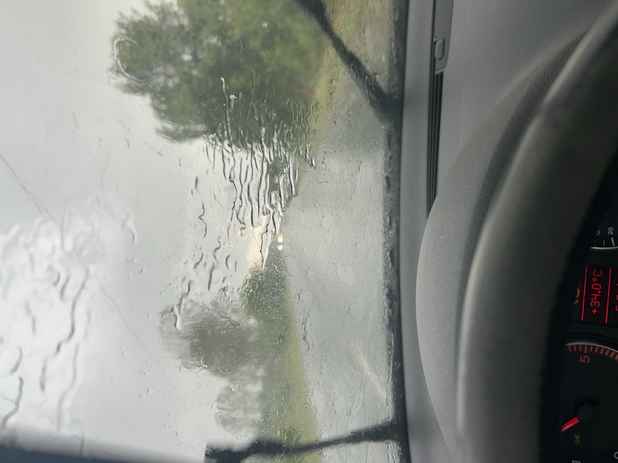 The image shows a view from inside a vehicle, likely a car, looking out through the windshield during a rainstorm. The windshield is covered with water droplets and streaks. Outside, trees and other vegetation are visible, obscured by the rain. The dashboard and steering wheel of the car are partially visible. The car's instrument panel displays a temperature of +34.0°C. The location is stated to be in Dunavtsi, Bulgaria.