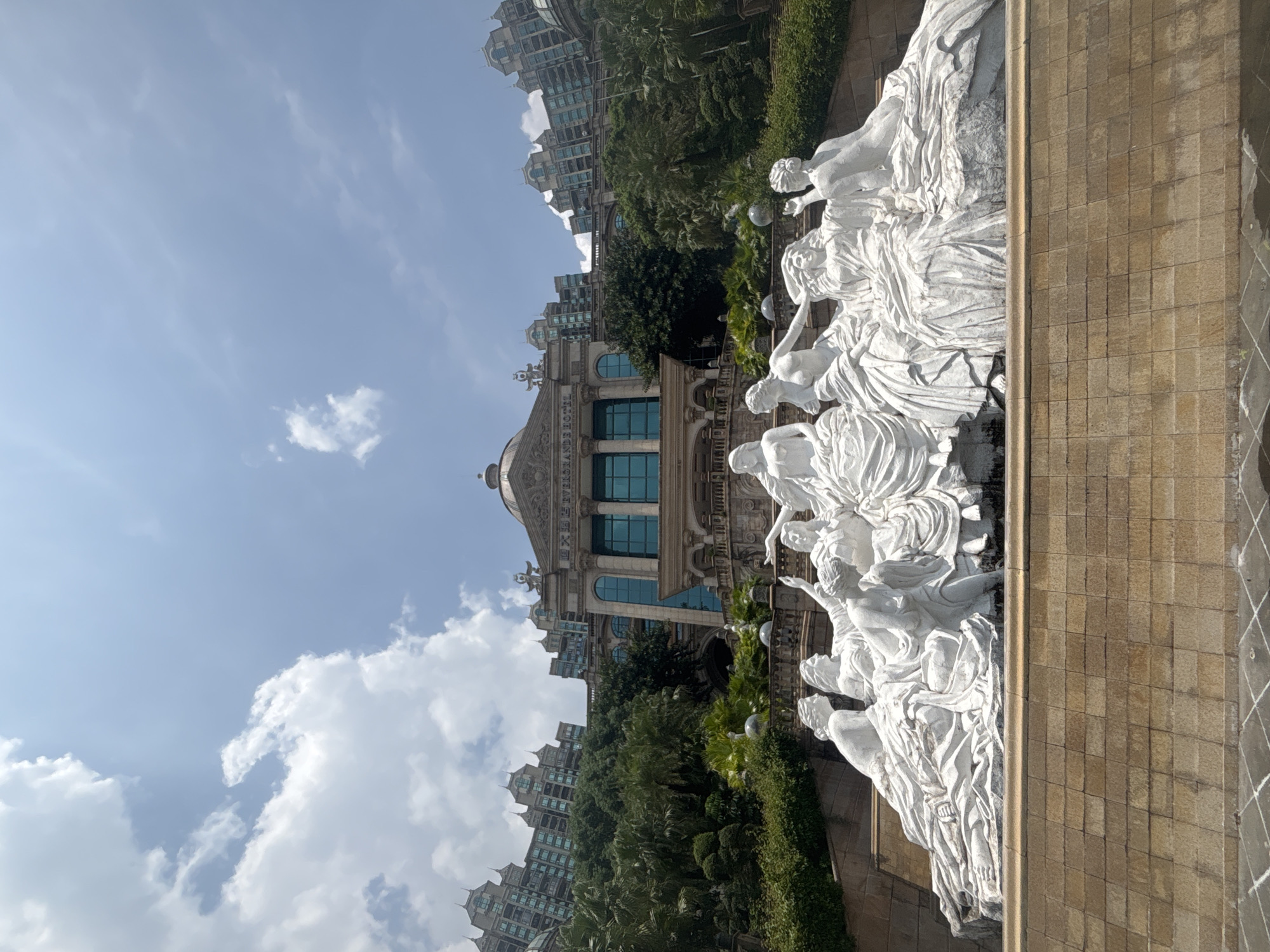 This is a low-angle, full shot of a white stone sculpture in front of a building, located in the Nanhai District of China. The sculpture depicts a group of women in flowing robes, some seated and some reclining, arranged in a semicircular formation. The building behind them is grand, with a domed roof, classical columns, and multiple stories with arched windows. Lush greenery and trees surround the building and slope upwards behind it. The sky is a clear blue with scattered white clouds. The foreground shows a textured stone wall and a body of water. The lighting suggests it is daytime.