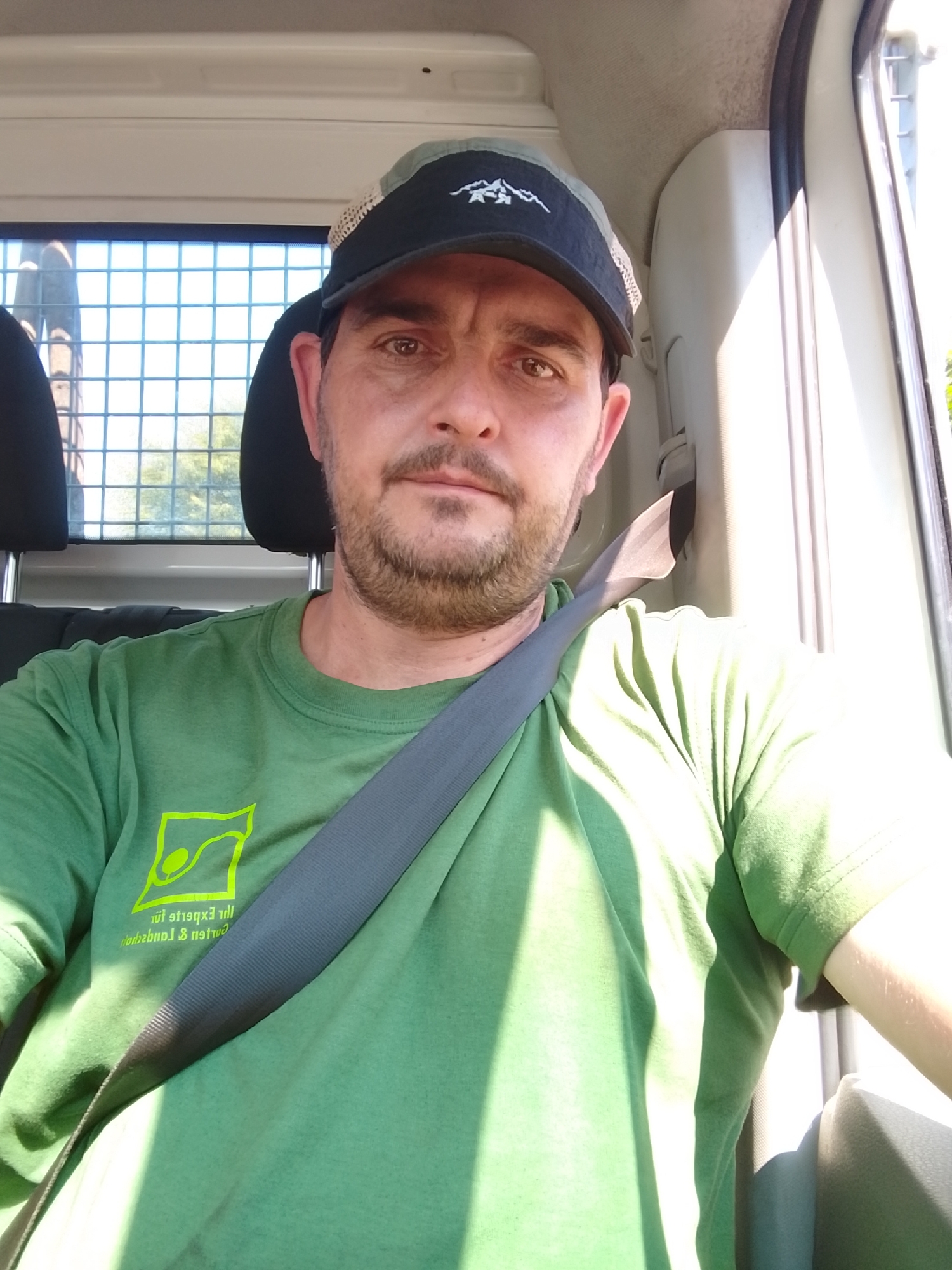 A man wearing a green t-shirt and a black and grey trucker hat is inside a vehicle, possibly a van, as indicated by the interior structure and the presence of a seatbelt. The man has a beard and appears to be looking directly at the camera, taking a selfie. The t-shirt has a bright yellow logo with text that is partially visible and appears to be in German, suggesting a local business or organization. The background shows the interior of the vehicle, with a grid-like window and the headrest of another seat. Sunlight is entering the vehicle from the right, creating strong shadows and highlights. The setting is not specifically identifiable as Schenefeld, Germany, based solely on visual cues.