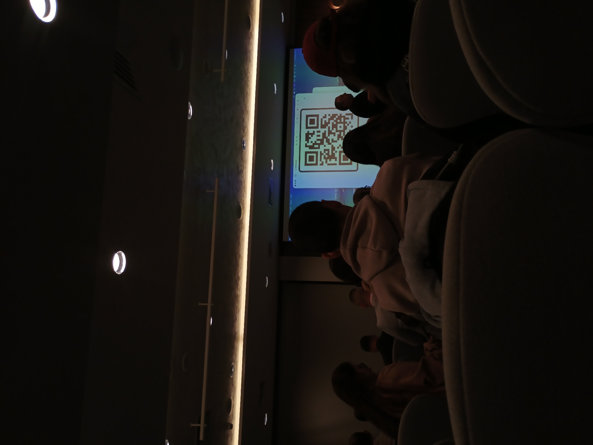 A dimly lit room in Plovdiv, Bulgaria, houses a presentation displaying a QR code on a screen, which is visible to an audience. The focus is on the screen and the backs of the heads of attendees seated in what appear to be cushioned chairs. The room has a row of evenly spaced recessed lights running vertically along the wall. The overall ambiance suggests a conference or training session.