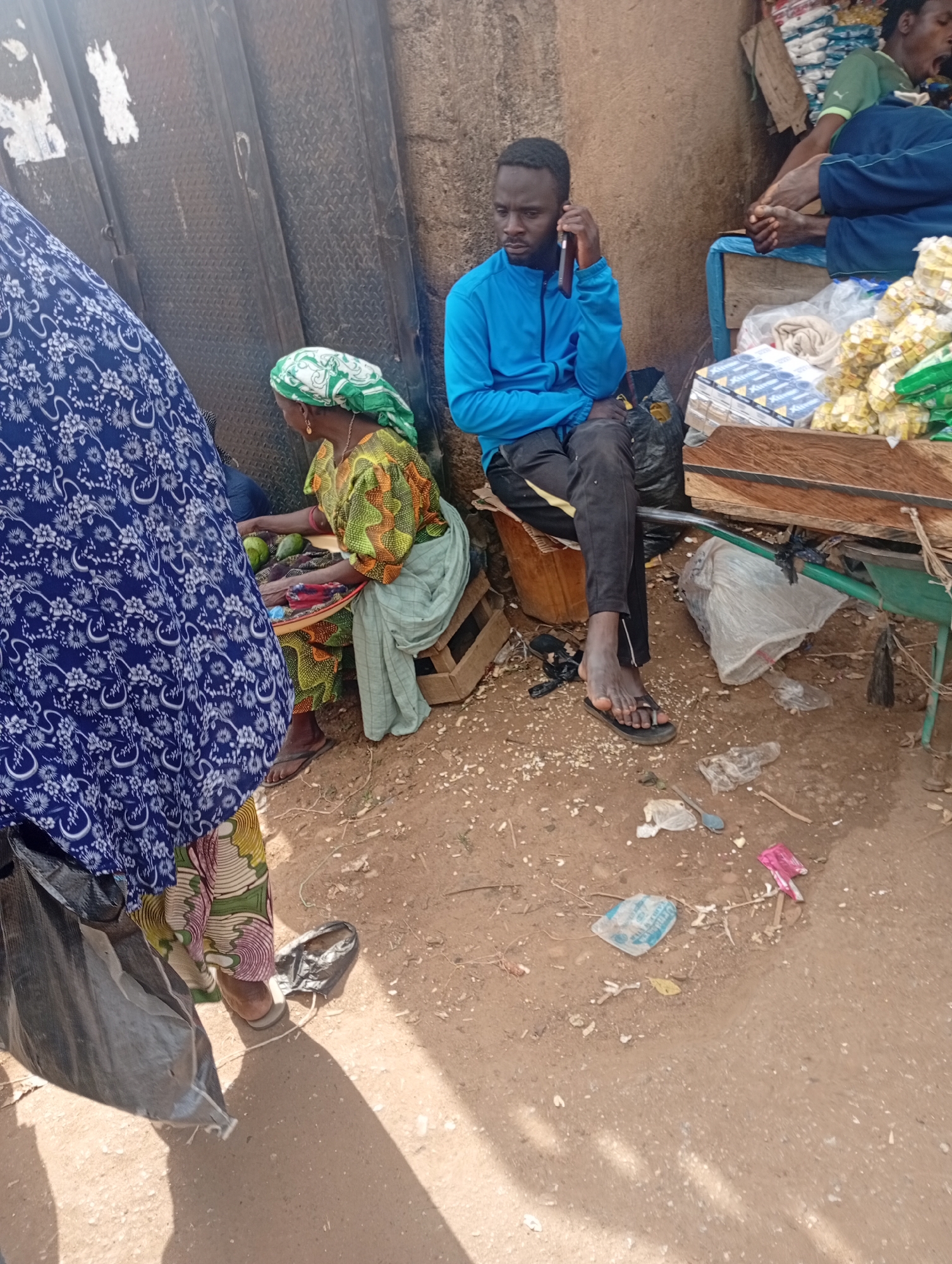 This image depicts a street scene in Jos, Nigeria, likely a market area. A young Black man in a bright blue jacket and black pants sits on a crate, talking on a mobile phone. To his left, an older Black woman wearing a green and white headscarf and a colorful patterned dress sits on the ground. She is tending to a tray of green fruits, possibly avocados, and a small pile of assorted items. A large blue and white patterned fabric covers a person on the far left, obscuring them from view. In the background, a metal grate or door is visible. The ground is dusty and littered with various debris, including plastic bags, wrappers, and small objects. A wooden stall or table laden with goods, some wrapped in plastic and others appearing to be packaged food items like bouillon cubes, is situated to the right and behind the seated individuals. The overall environment suggests an outdoor vendor setting in a bustling urban area.