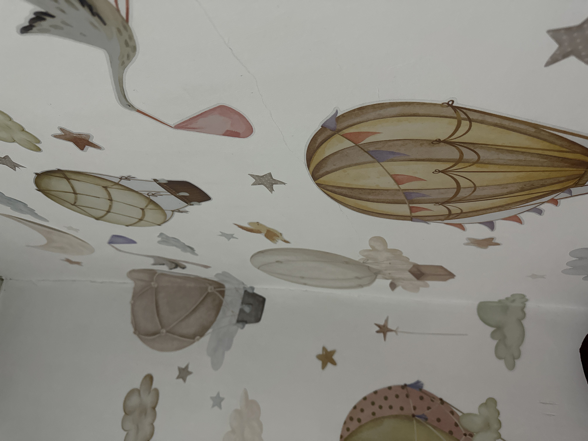 The image is a close-up, eye-level, interior shot of a wall in a room, possibly a nursery. The wall is decorated with watercolor-style decals of hot air balloons, clouds, and a stork carrying a bundle. The color palette consists of muted tones of brown, beige, yellow, pink, and gray. The overall aesthetic is whimsical and childlike. The room appears to be located in Тайгараев, Kyrgyzstan, although there are no specific architectural details visible to confirm this location beyond the provided prompt. The lighting is natural, casting soft shadows on the wall. There are no people or animals present in the image.