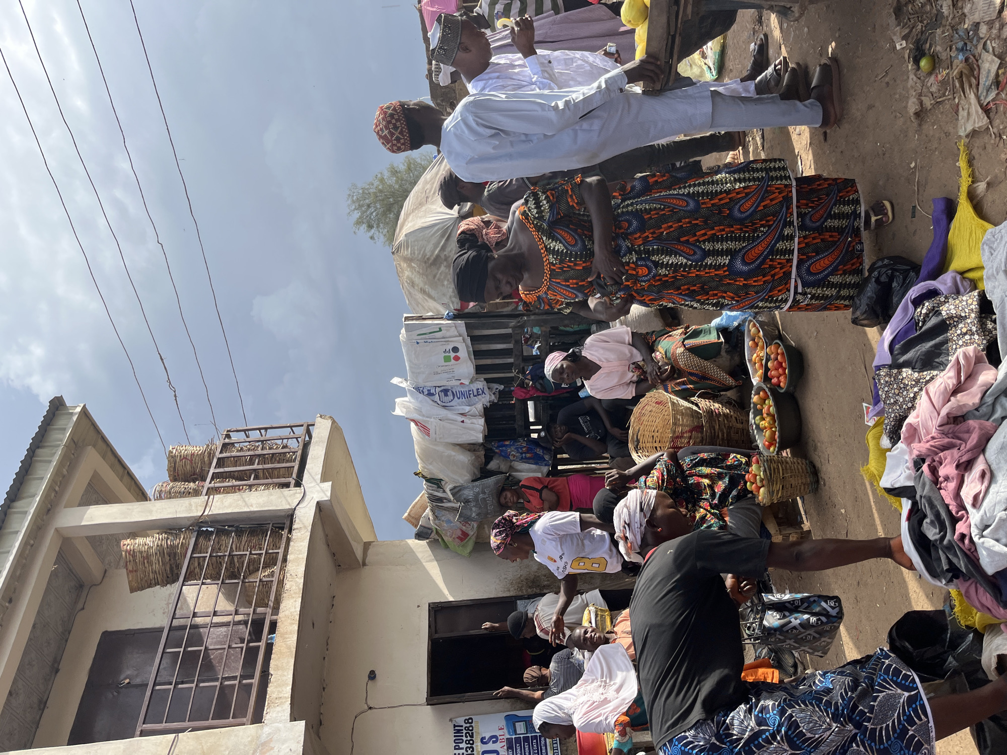 The image depicts a busy outdoor market scene in Rafin Par, Nigeria. The foreground features a person in a black shirt and patterned blue and white skirt bending over a pile of colorful clothing on the ground. To the right, a wooden table is laden with a crate of yellow fruit, possibly lemons or gourds, and a person in a white robe and red patterned cap stands next to it. In the mid-ground, a woman in a vibrant orange, blue, and black patterned dress is the focal point, appearing to be engaged in conversation. Other people are scattered throughout the scene, some dressed in traditional attire, others in casual clothing. Behind the people, stalls are set up with various goods, including baskets stacked on a balcony above, sacks, and more produce. The environment is dusty and unpaved, characteristic of a market in a Nigerian city. The sky above is overcast with some patches of sunlight breaking through. The overall impression is one of lively commerce and daily life in a community.