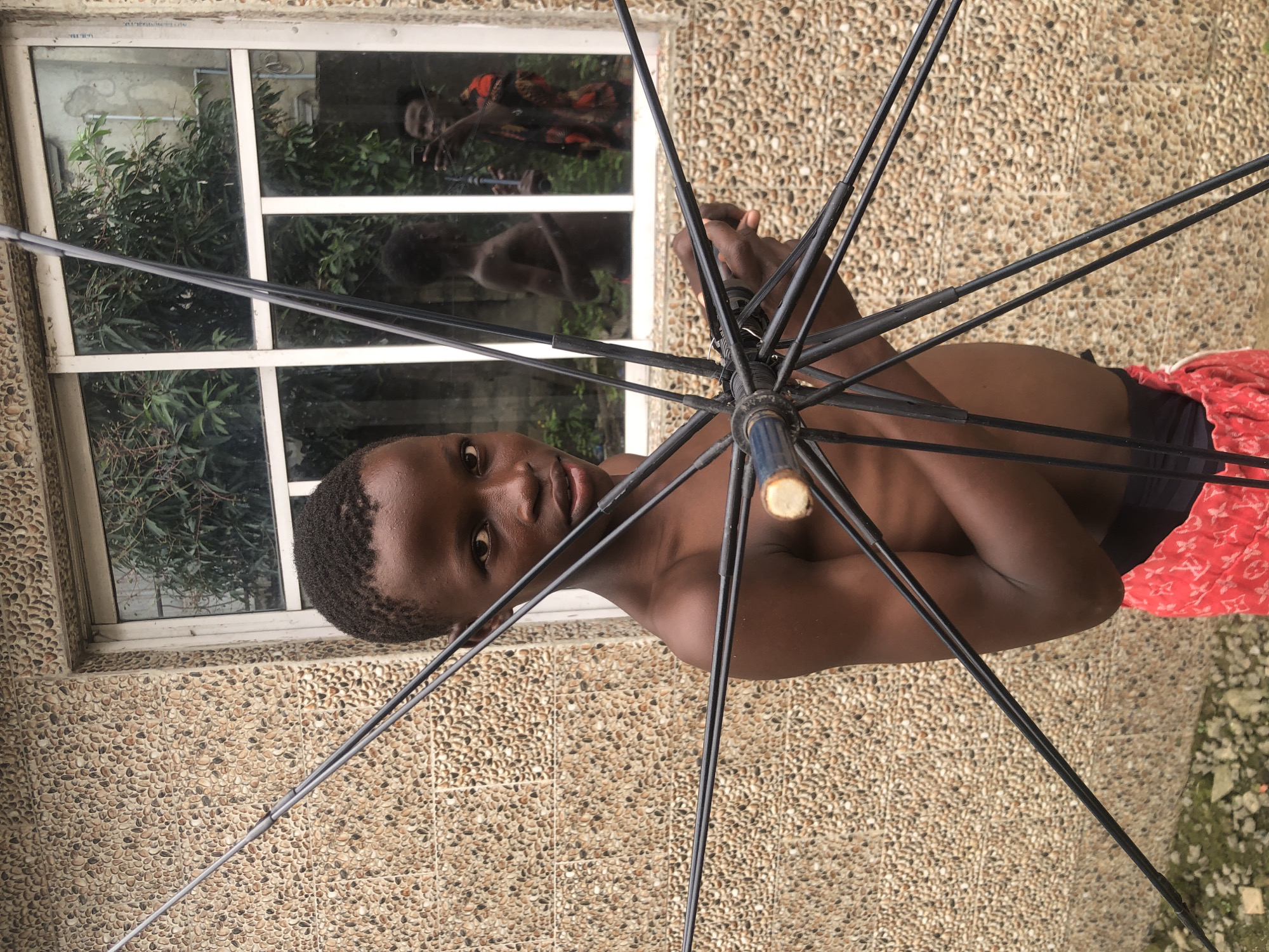 In Ado, Nigeria, a young Black male stands shirtless in front of a pebble-clad building wall. He is holding the spokes of a black umbrella, which are splayed outwards. A window with multiple panes is visible behind him, reflecting trees and two other figures. One reflected figure appears to be a young Black male, shirtless, and another is a Black male holding a phone, possibly taking a selfie. The young male in the foreground wears red patterned shorts. The ground is covered with gravel and some green vegetation. The lighting suggests an overcast day or shaded area.