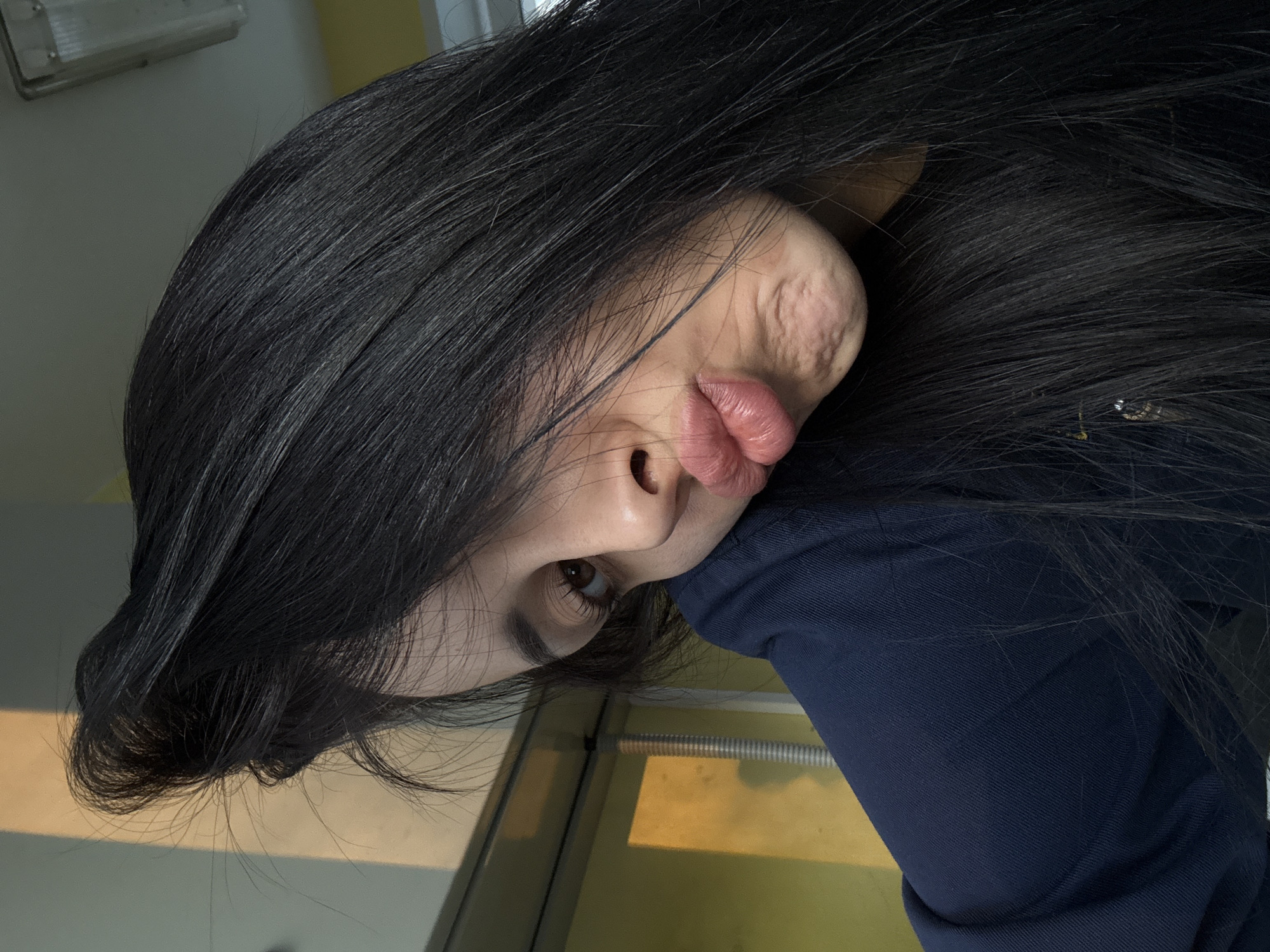 A selfie-style photograph shows a person with long, black hair. The person is wearing a dark blue garment. The photograph is taken at an angle, with the person's face tilted. The background appears to be an interior space with yellow and light-colored surfaces. It is not possible to determine the location as Molodezhnyi, Russia from the image content.