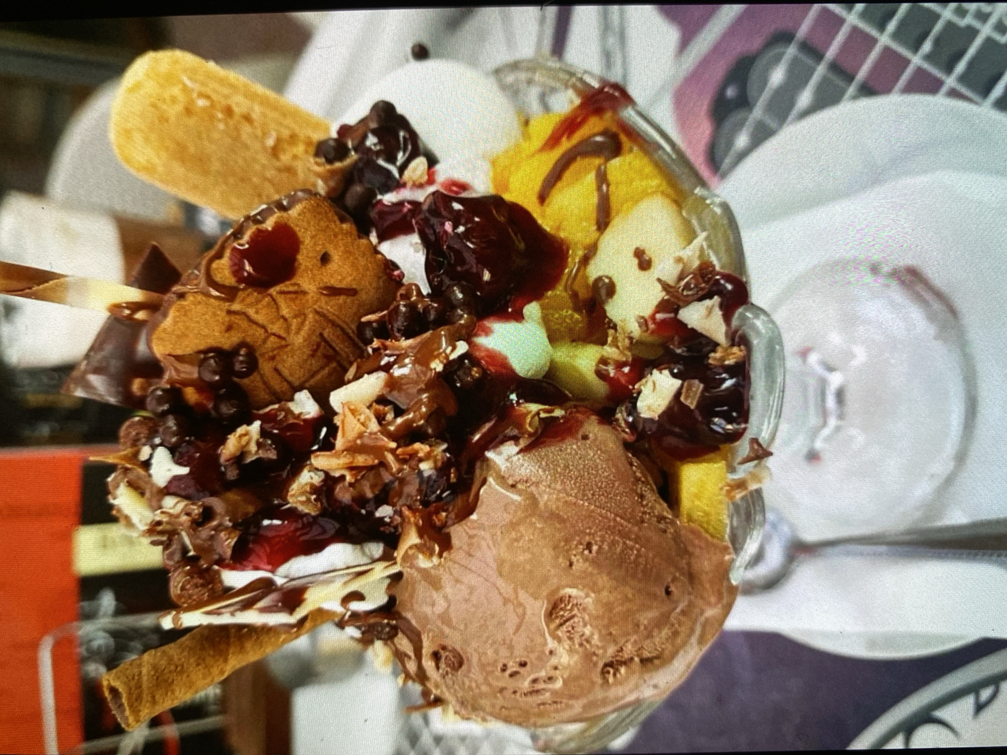 The image captures a vibrant and elaborately presented ice cream sundae in a clear glass dish, viewed from a slightly elevated, tilted angle. No people are visible.
The sundae features multiple scoops of ice cream, including a rich chocolate flavor, a creamy white (likely vanilla or similar), and a bright yellow (possibly a fruit flavor like mango or pineapple). It is generously topped with a medley of ingredients: dark red cherry or berry compote, glossy chocolate syrup, chocolate chips, and shredded nuts or coconut flakes, which add texture and visual appeal. Small green fruity bits are also visible.
Several decorative biscuits and wafers are incorporated into the dessert. A long, rectangular golden-brown wafer or ladyfinger stands tall at the back. A patterned cookie, possibly heart-shaped or star-shaped, is partially submerged and drizzled with chocolate syrup. A rolled wafer stick is positioned towards the front. A glass spoon or straw is also visible within the sundae, ready for use.
The sundae rests on a light-colored, possibly white, table surface. To the right of the glass, a silver spoon and the edge of a white plate or bowl are faintly visible. The background is softly blurred, revealing hints of a reddish-orange object on the left and a white, grid-like pattern, possibly furniture, on the right. The setting appears to be indoors, likely a cafe or restaurant, with bright, possibly artificial, lighting. There is no visible text in the image.