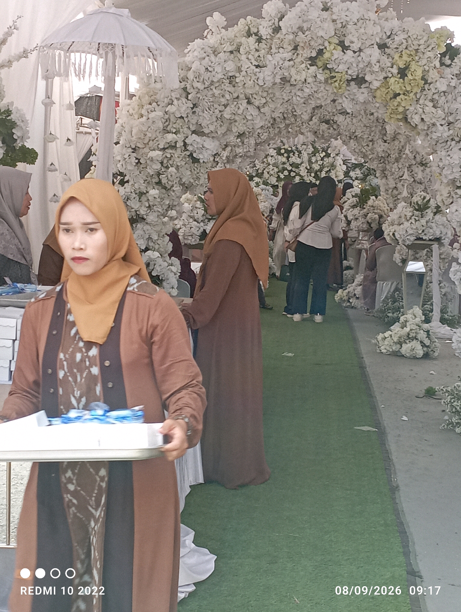 The image captures a gathering with a significant floral display, likely a wedding or similar event, set outdoors. In the foreground, a woman wearing a brown hijab and a brown dress with a patterned undergarment is holding a white tray with blue ribbon. Her expression appears serious as she looks to her left. Behind her, another woman in a brown hijab is visible, along with other individuals in the background, some of whom are wearing white shirts and dark pants. The environment features an artificial green carpet covering the ground, and an abundance of white flowers forming arches and decorative elements. The backdrop suggests a tented structure, indicated by the white fabric and a decorative white umbrella-like object. The image is dated "08/09/2026 09:17" and tagged "REDMI 10 2022". The location is identified as Dukuhseti, Indonesia.