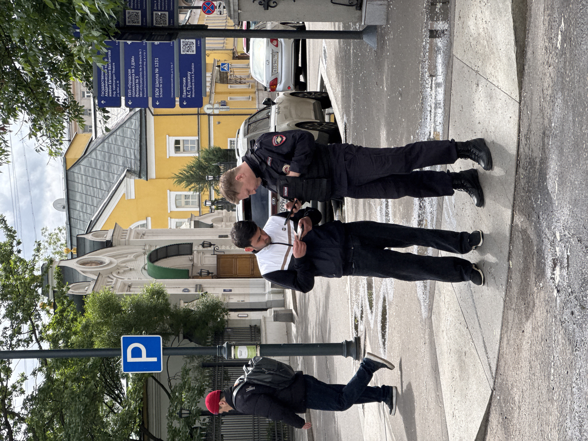 The image shows an outdoor scene on a city street, likely in Moscow, Russia. Two men are standing together, looking at their phones. One is in civilian clothing, the other is a police officer wearing a black uniform with "полиция" (police) written on it. A third man wearing a red beanie, dark jacket, and backpack is walking away from the group. Behind them, there is a yellow building and a street sign indicating directions to locations such as the "Monument to A.S. Pushkin" and "Eug. Vakhtangov Theatre", identifiable through the Russian Cyrillic script and names of cultural landmarks associated with Moscow. A blue parking sign "P" is visible on the left. The pavement is wet, suggesting it recently rained. A KIA car can be seen parked behind the group.