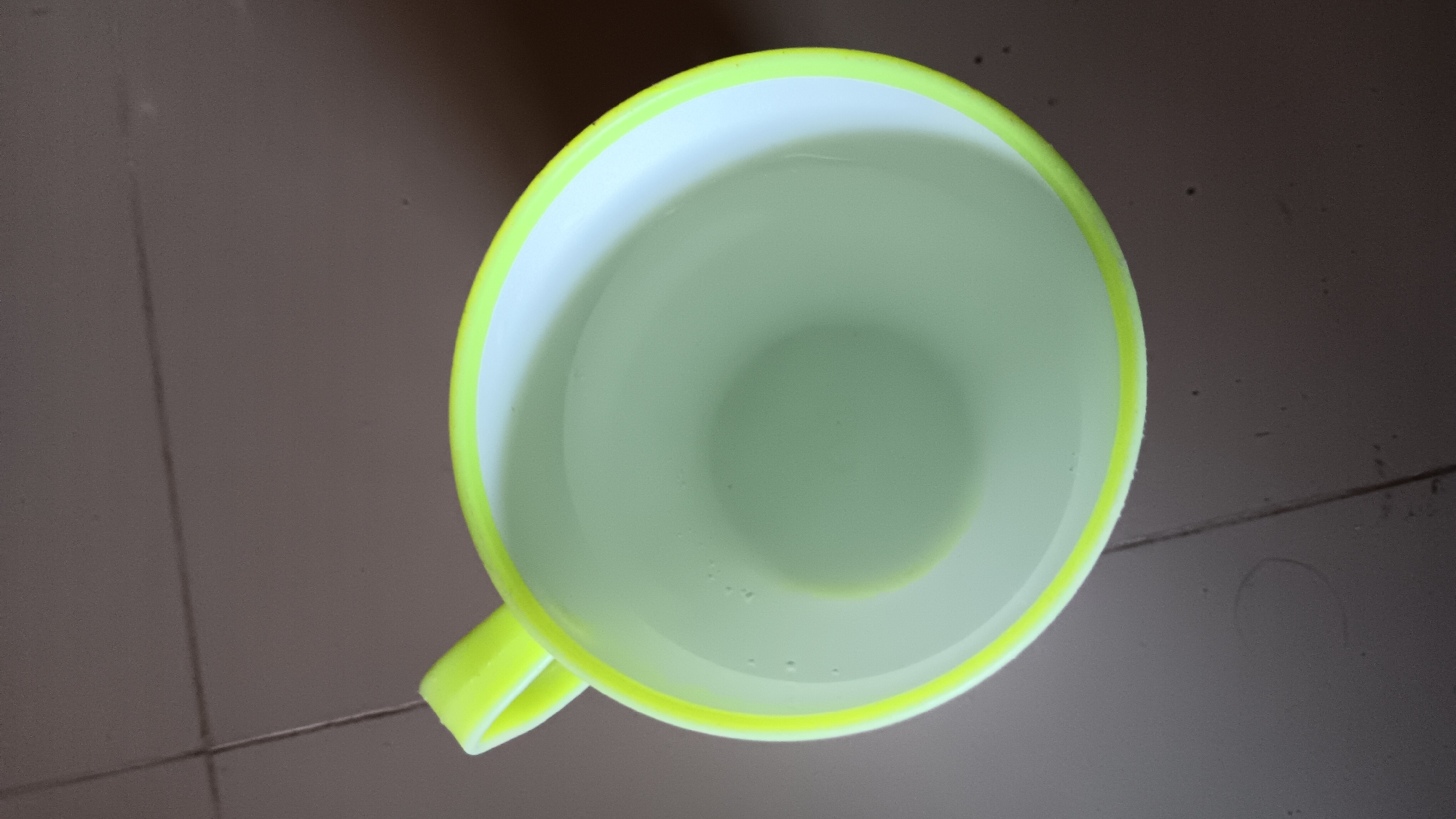 A top-down, slightly angled view shows a bright yellow-handled mug filled with clear liquid, possibly water, resting on a brown tiled surface. The mug's rim is also outlined in the same bright yellow. The liquid within the mug has a subtle green tint and appears to be partially filled, with some small bubbles visible on the surface. The brown tiles have visible grout lines and appear to be slightly dusty or worn in places. The lighting suggests an indoor environment, possibly a home or simple office setting. No people or animals are present in the image. The scene is static and devoid of any action. The location is identified as Sonobekel, Indonesia.