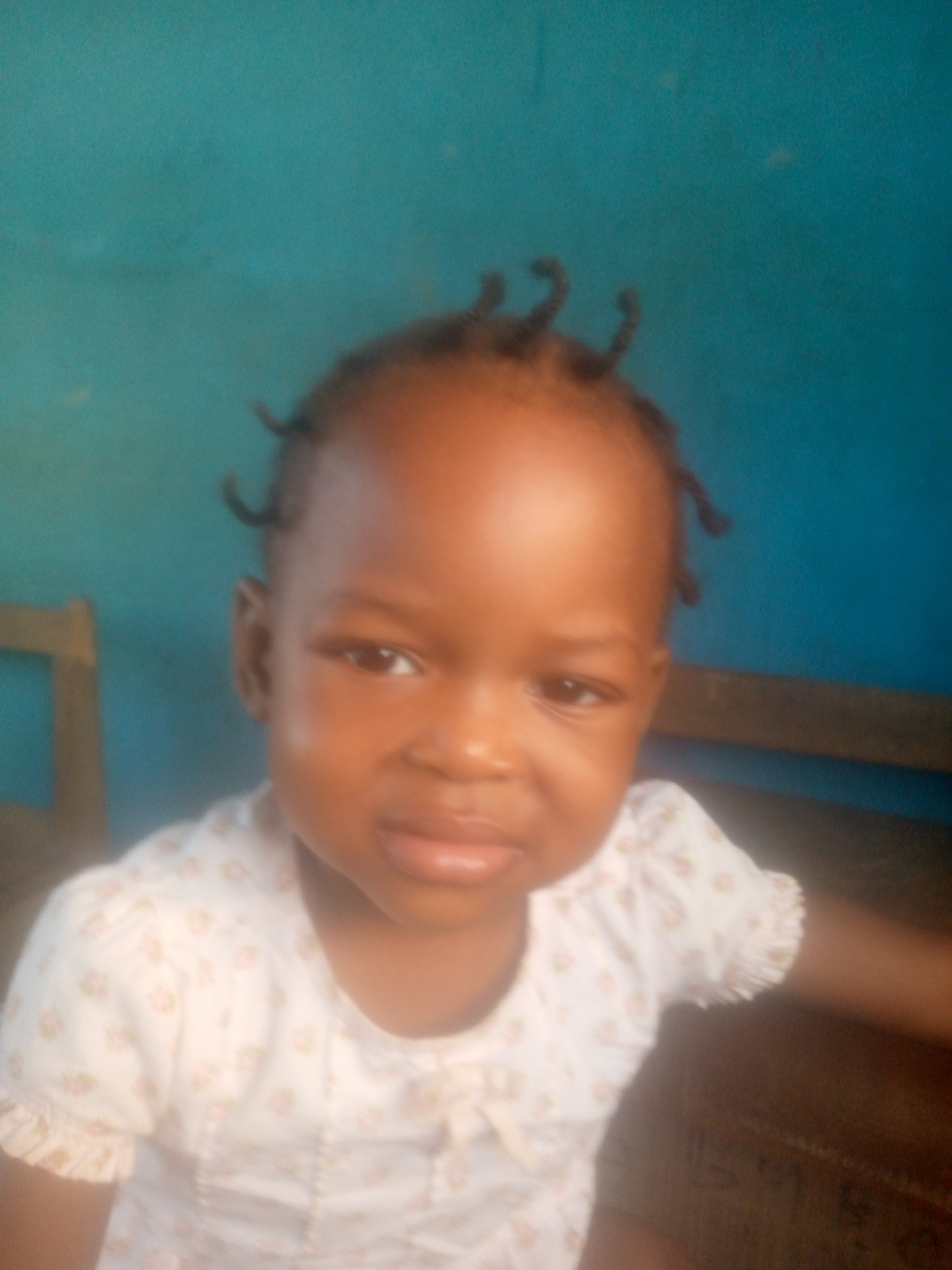 A young child with dark skin and short, curled hair stands in front of a teal wall. The child wears a light-colored, short-sleeved shirt with small floral patterns. The location is Unknown, Unknown.