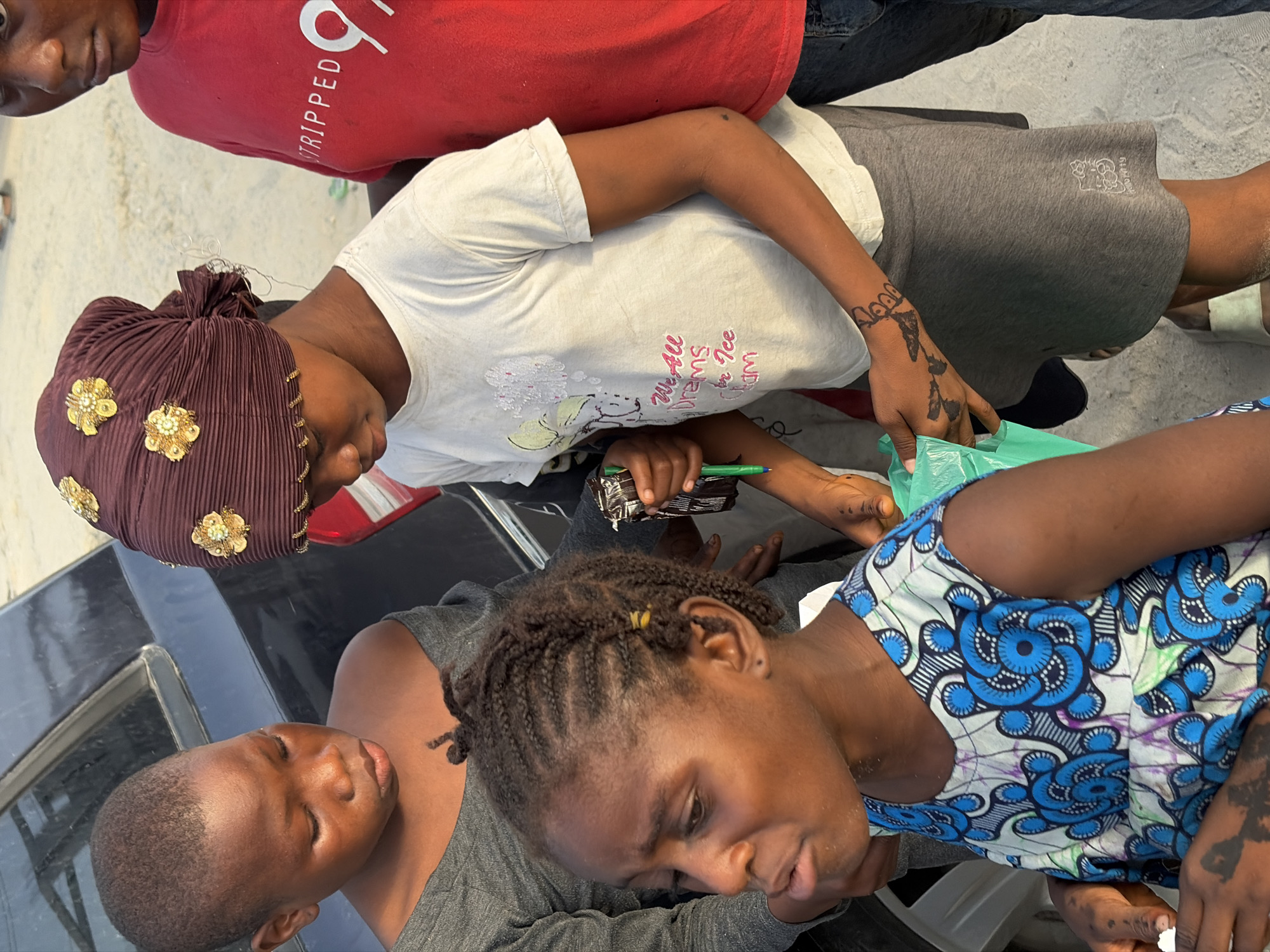 The image shows a group of people outdoors, likely in Alaguntan, Nigeria. At least four individuals are partially visible. One wears a red shirt with white text, and another wears a white t-shirt and grey shorts, with temporary tattoos on the hands. A third person is wearing a brown head covering adorned with gold floral accents. Another individual is wearing a blue and white patterned dress. The group appears to be interacting, with one person holding a pen and some packaged goods. The scene is situated on and around the trunk of a black car. The environment has a daytime, outdoor setting.