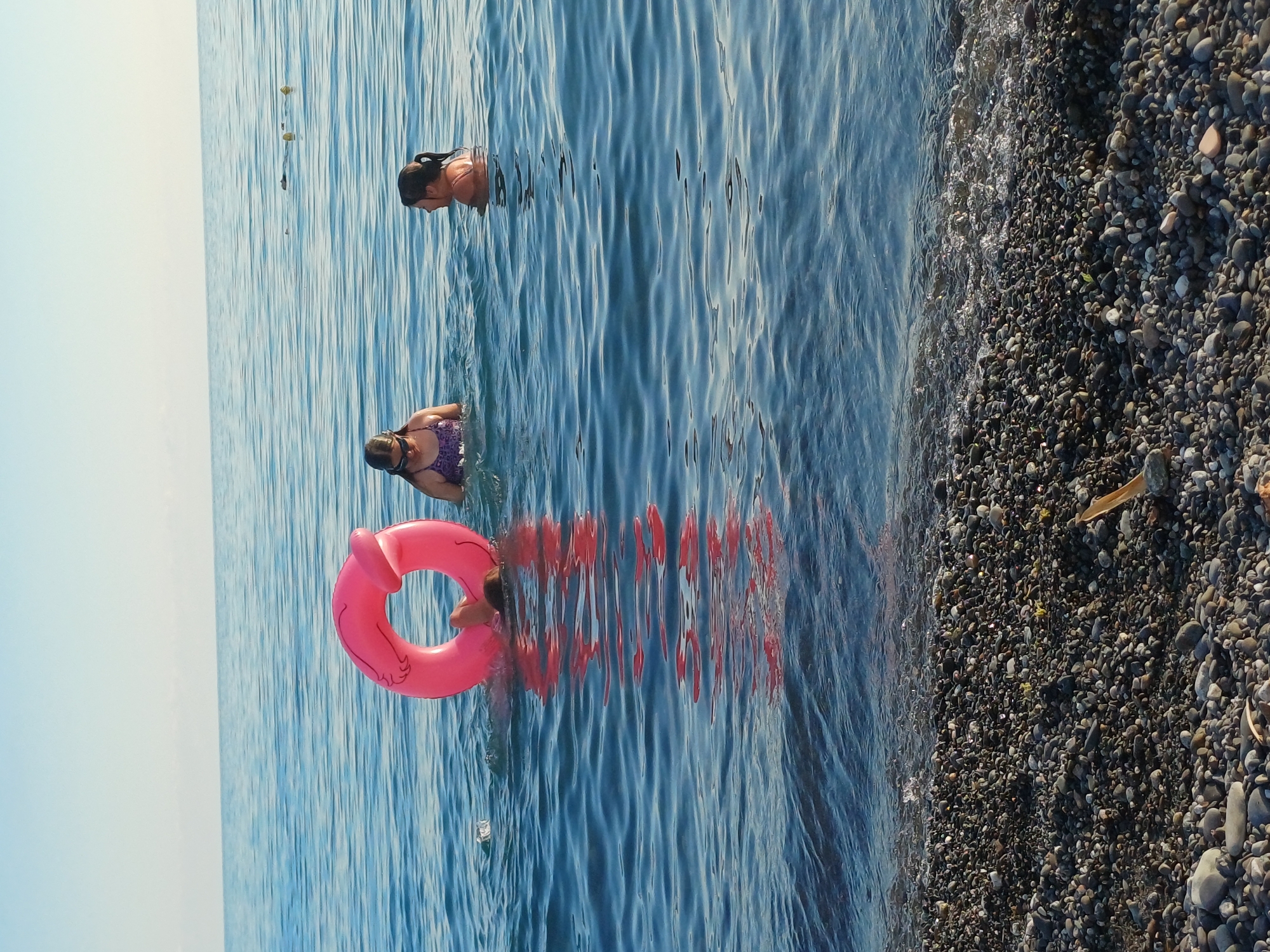 The image depicts a serene beach scene in Sochi, Russia, with three individuals enjoying the calm waters of the Black Sea. In the foreground, a pebbled shoreline meets the gentle waves. Two girls are visible in the water: one, wearing a purple swimsuit and goggles, is partially submerged, while another, with dark hair pulled back, is looking to the side, her shoulders and head above the water. A pink inflatable ring floats in the water, with one person's head and shoulders visible inside it, suggesting playful activity. The sea is a tranquil blue, stretching to a hazy horizon under a clear sky, indicative of a pleasant day in the popular Black Sea resort city.