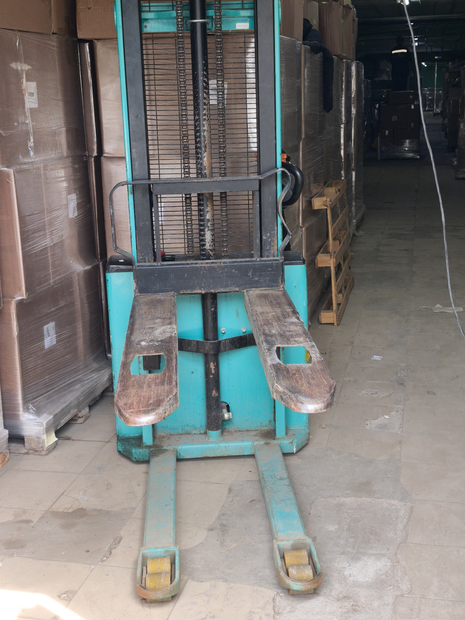 A turquoise and black industrial forklift is situated indoors in what appears to be a warehouse or storage facility. The forks of the forklift are brown with rectangular holes. The machine is parked on a grey, tiled floor. Adjacent to the forklift are stacked boxes, some wrapped in clear plastic, and wooden pallets. Additional boxes are visible further down the aisle. A light fixture hangs from the ceiling in the background. There are no people visible in close proximity to the forklift.