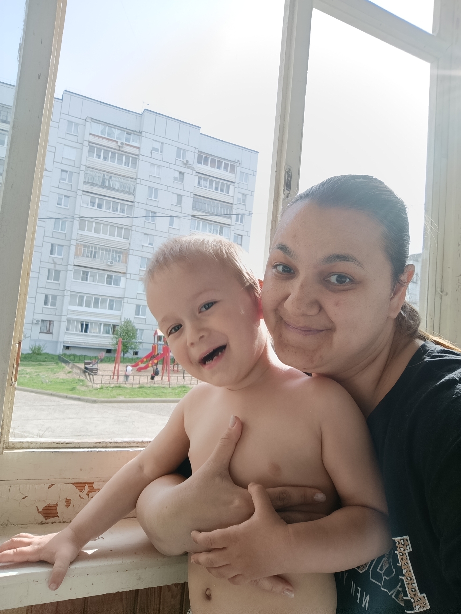 A woman and a young child are captured in a close-up indoor shot, likely a selfie, with a backdrop of a residential building and a playground. The child, shirtless, is held by the woman, who is wearing a dark-colored shirt with some visible text. Both are looking at the camera. The setting appears to be near a window with a view of a residential area. The playground in the background has red and blue structures. Based on the building style and other details, the location is consistent with Chaikovsky, Russia.