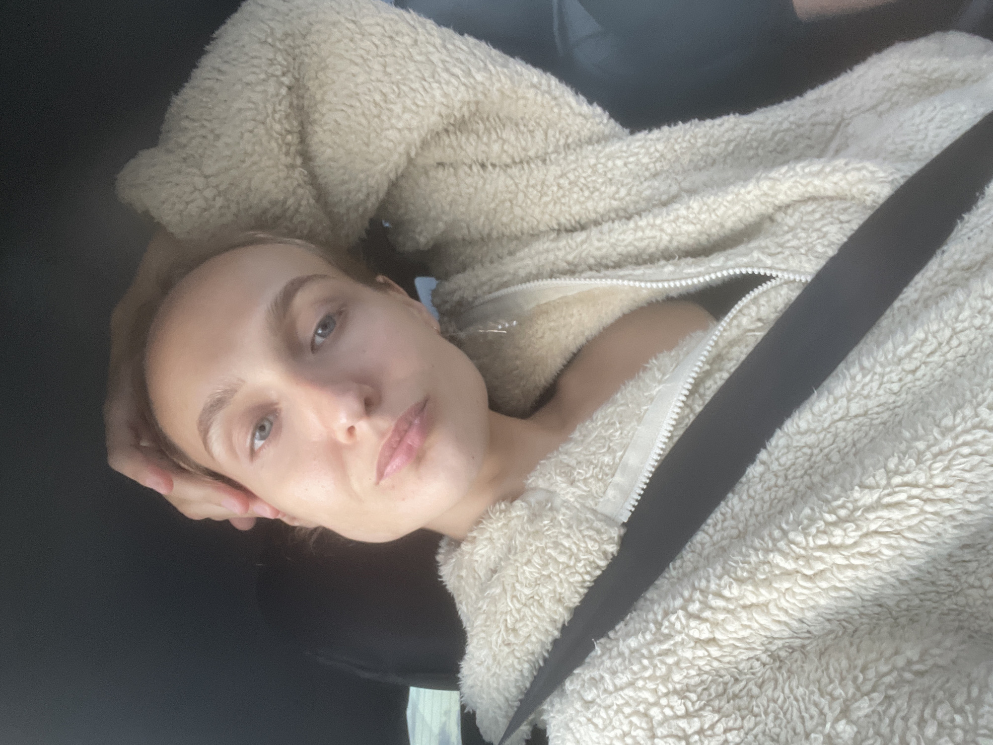 A woman is in the driver's seat of a car, wearing a fluffy cream-colored jacket with a zipper. A black seatbelt is across her chest. Her right hand is on the back of her head, and her left arm is bent, resting on her lap. The background is dark, suggesting the interior of the car. The image appears to be a selfie. The location is Pirogovo, Russia.