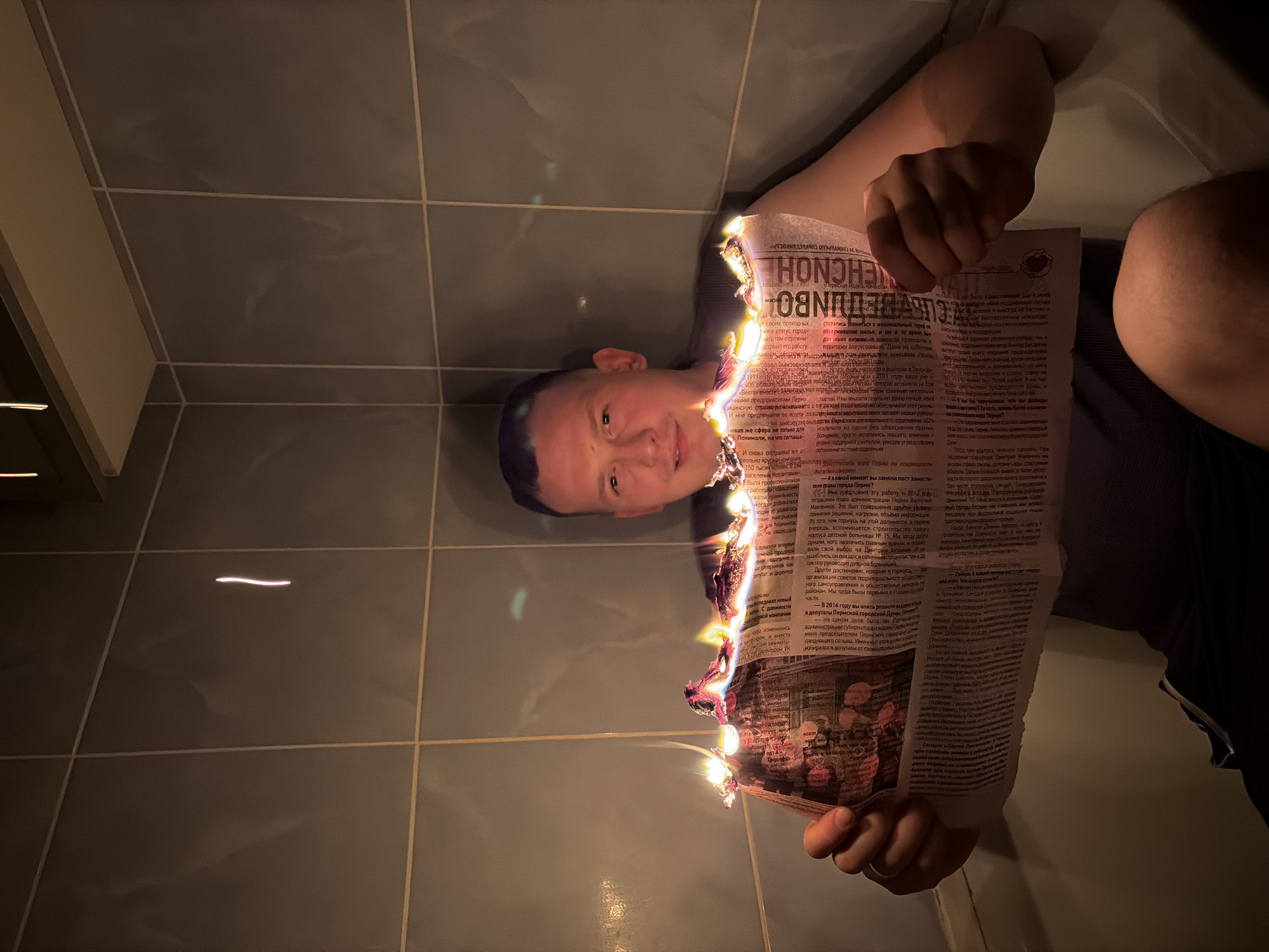 The image depicts a man in what appears to be a bathroom, seated on the edge of a bathtub. He is holding a newspaper that is actively burning along its top edge. The flames are bright orange and yellow, with some smoke rising from the paper. The man is looking directly at the camera with a slight smile. The bathroom walls are tiled in a muted gray color, and there is a shelf above the man's head. The lighting is dim, with the primary illumination coming from the burning newspaper, casting a warm glow on the man's face and the surrounding tiles. There is no indication of the city of Chaikovsky, Russia, in the image itself, and therefore no location-specific details can be inferred from the visual content.