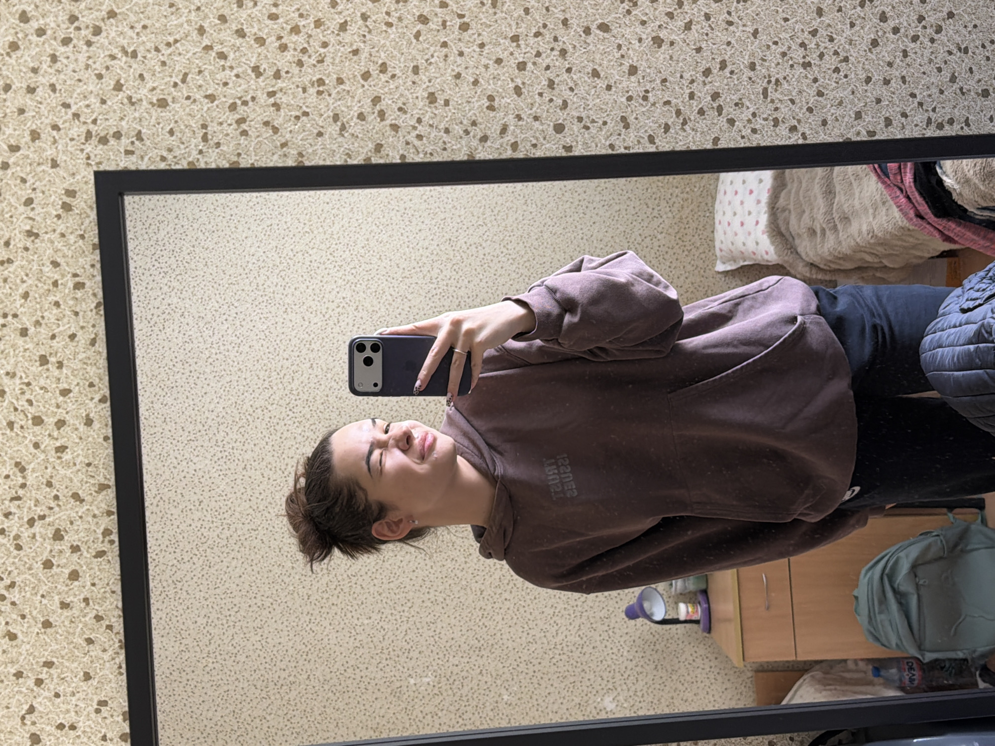 A young woman is captured in a mirror selfie, taken indoors in what appears to be a bedroom or living area in Sofia, Bulgaria. She stands in front of a large, black-framed rectangular mirror, holding a dark-colored smartphone with a patterned case to take the photo. Her hair is pulled back, and she wears a brown or mauve hooded sweatshirt and dark trousers. She has a playful expression, with her eyes partially closed and a slight smile. The text "TRUST ISSUES" is visibly reversed on her sweatshirt.
Reflected in the mirror, behind the woman, are elements of the room. A bed or couch with blankets and pillows is visible to the upper right. To the lower right, a light wooden cabinet holds a purple and white lamp. A light green/grey backpack and a plastic water bottle with the reversed partial text "DEAR" on its label are on the floor next to the cabinet. The wall visible outside the mirror and in the reflection features a distinctive beige wallpaper with small brown, speckled patterns. The overall lighting suggests it is daytime indoors.