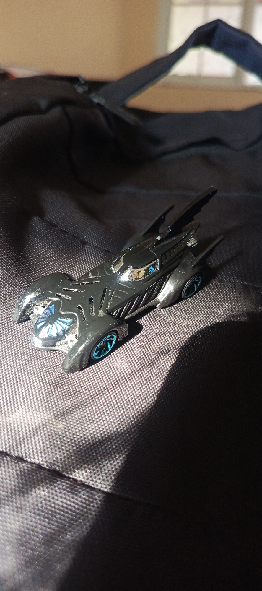 An overhead close-up shot shows a black toy Batmobile resting on a black textured fabric surface. The toy car features blue accents on its wheels and front grille. The background consists of a beige wall and a window, with a dark shadow cast over the right side of the image. The scene is indoors. The location is not specified.