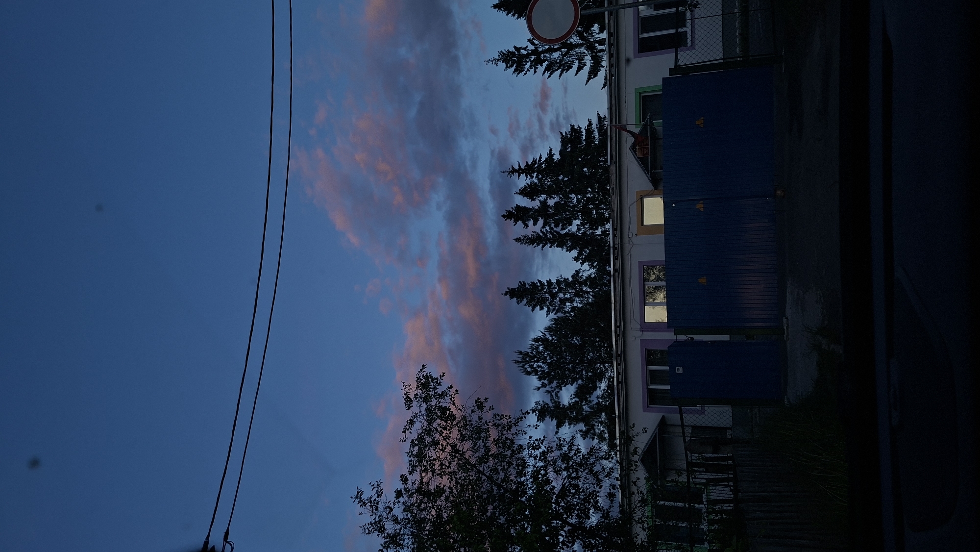 The image depicts a twilight scene, likely in Забалоцкі сельскі Савет, Belarus, as suggested. The sky is a mix of deep blue and pink-tinged clouds. Power lines run across the left portion of the frame. To the right of the sky, there are silhouettes of trees, including some evergreen trees. Adjacent to the trees is a building with lighted windows, indicating occupancy. In front of the building, there are blue structures, possibly storage containers. A road sign is also visible above the trees. The overall setting suggests a quiet residential area at dusk.