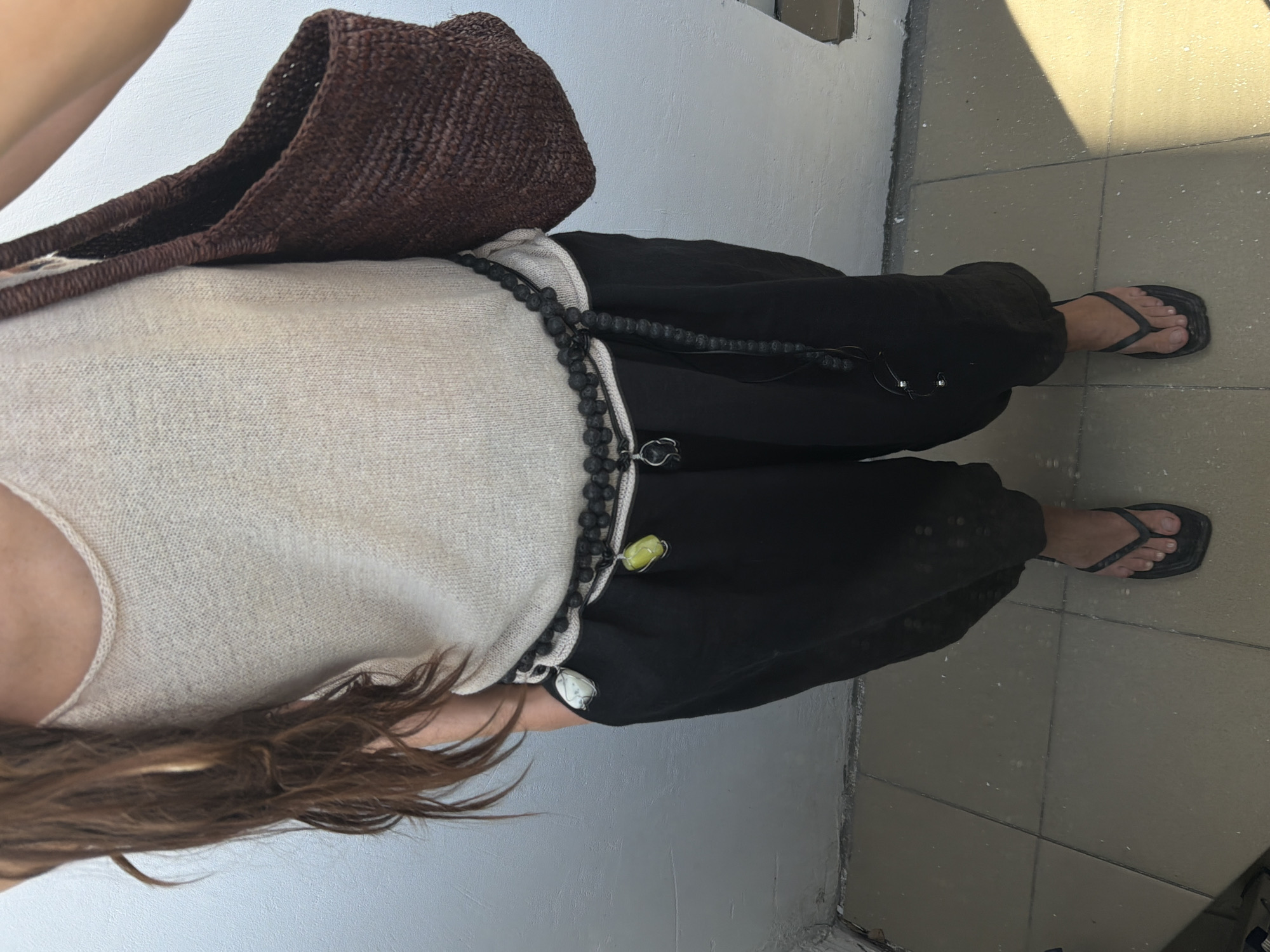 A medium shot shows a person from the mid-torso down, standing outdoors in daylight. The person is wearing a light-colored knit tank top and loose black pants that end above the ankles. A dark woven bag is slung over the right shoulder, and a beaded necklace, possibly a belt, with several charms hangs around the waist. The person is wearing black thong sandals. The background consists of a white wall and a tiled floor. The scene appears to be on a balcony or patio. There is no indication of the location being in Moscow, Russia.