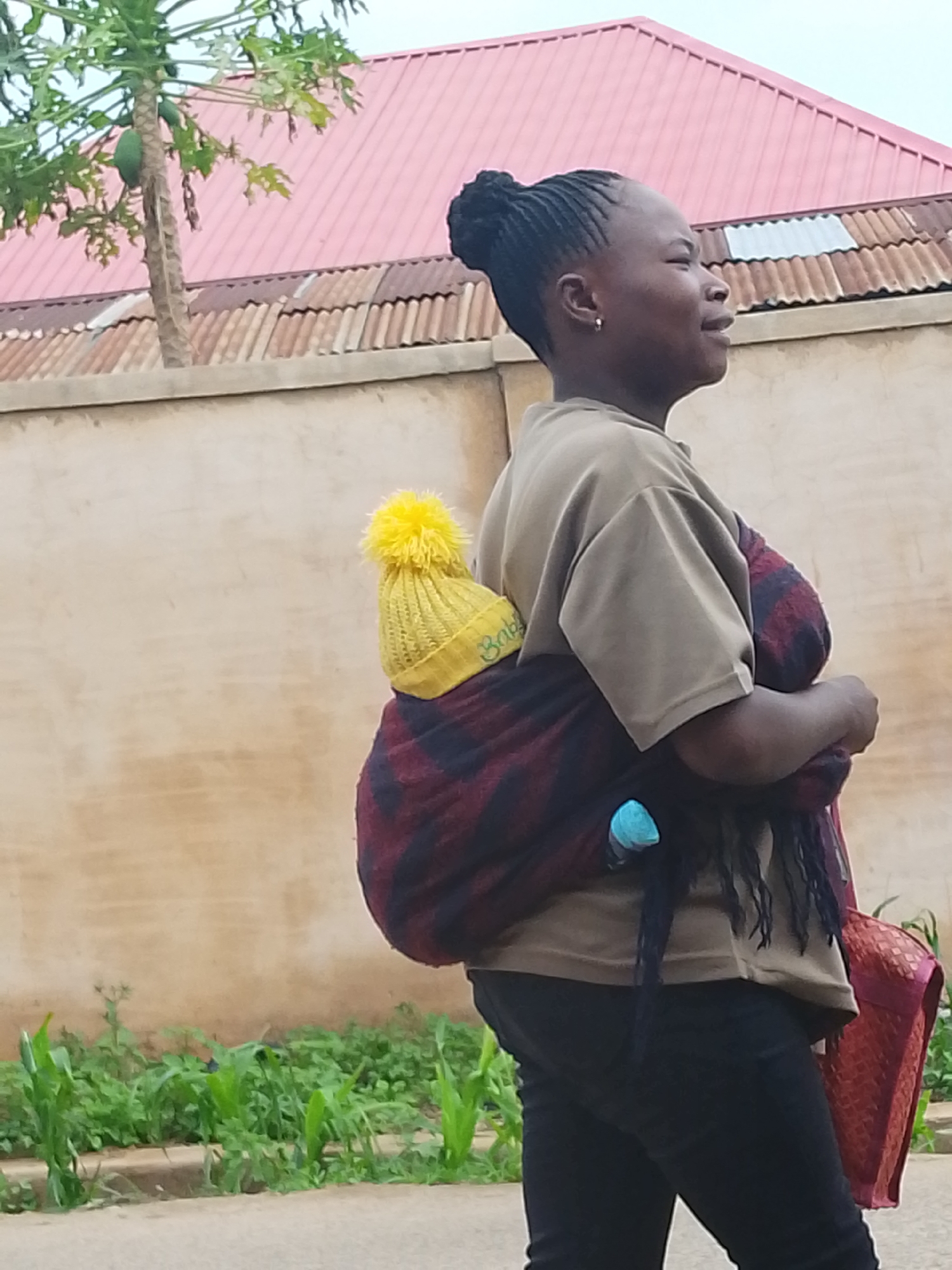 A side-view medium shot shows a woman in Jos, Nigeria, walking along a sidewalk. She has dark skin and hair styled in cornrows gathered into a bun. She wears a light brown t-shirt and dark pants. A patterned cloth wraps around her back, supporting what appears to be a baby wearing a yellow hat with a pom-pom on top. A blue bottle is visible in the cloth wrapping. The woman carries a woven red bag in her hand.
Behind her is a light-colored wall topped with corrugated metal. A red roof can be seen behind the wall, along with a small papaya tree. Green plants grow along the edge of the sidewalk.