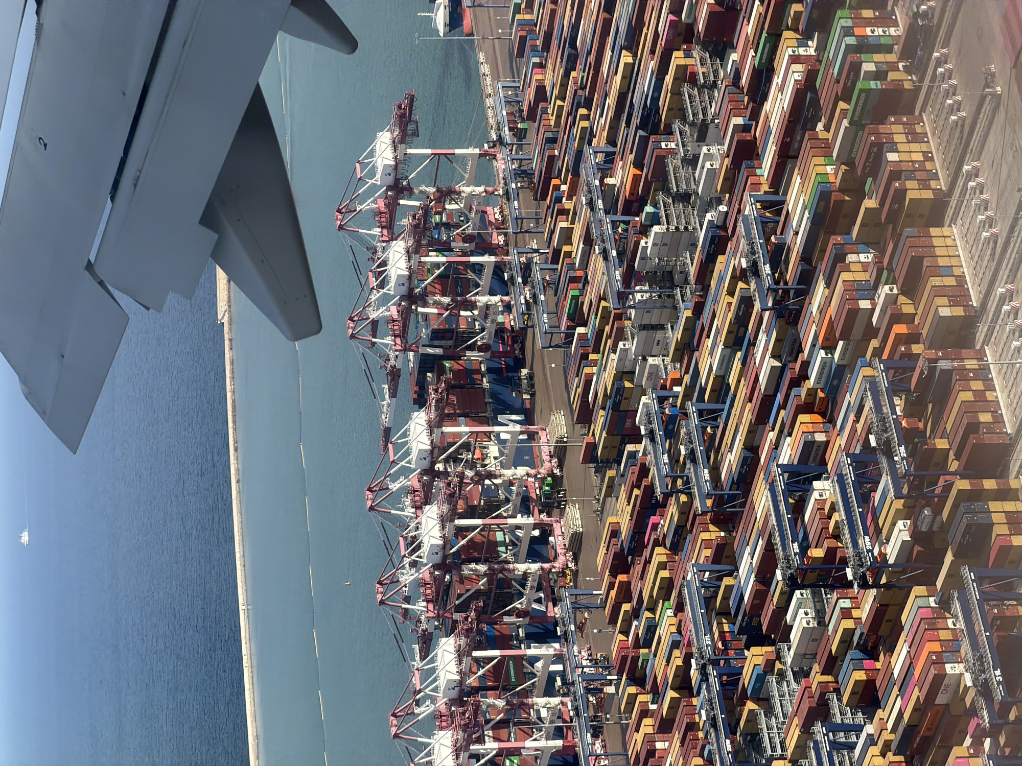 The image shows an aerial view of a large container port with numerous shipping containers stacked high and organized in rows. Several massive gantry cranes, painted in pink and white, are positioned over the containers, suggesting active loading and unloading operations. The port is adjacent to a body of blue water, with a breakwater or seawall separating it from a wider expanse of the ocean where a small white ship is visible in the distance. An airplane's wing, a portion of the fuselage, and part of the flap system are prominently in the foreground, indicating the photo was taken from an aircraft in flight. The scene is lit by daylight, casting shadows consistent with an overhead sun. The overall impression is one of a busy, industrial maritime hub. While the prompt specifies Barcelona, Spain, there are no discernible landmarks or text within the image to confirm this location. The scene is comprised solely of the port infrastructure, containers, water, and the aircraft wing. There are no people visible in the image.
