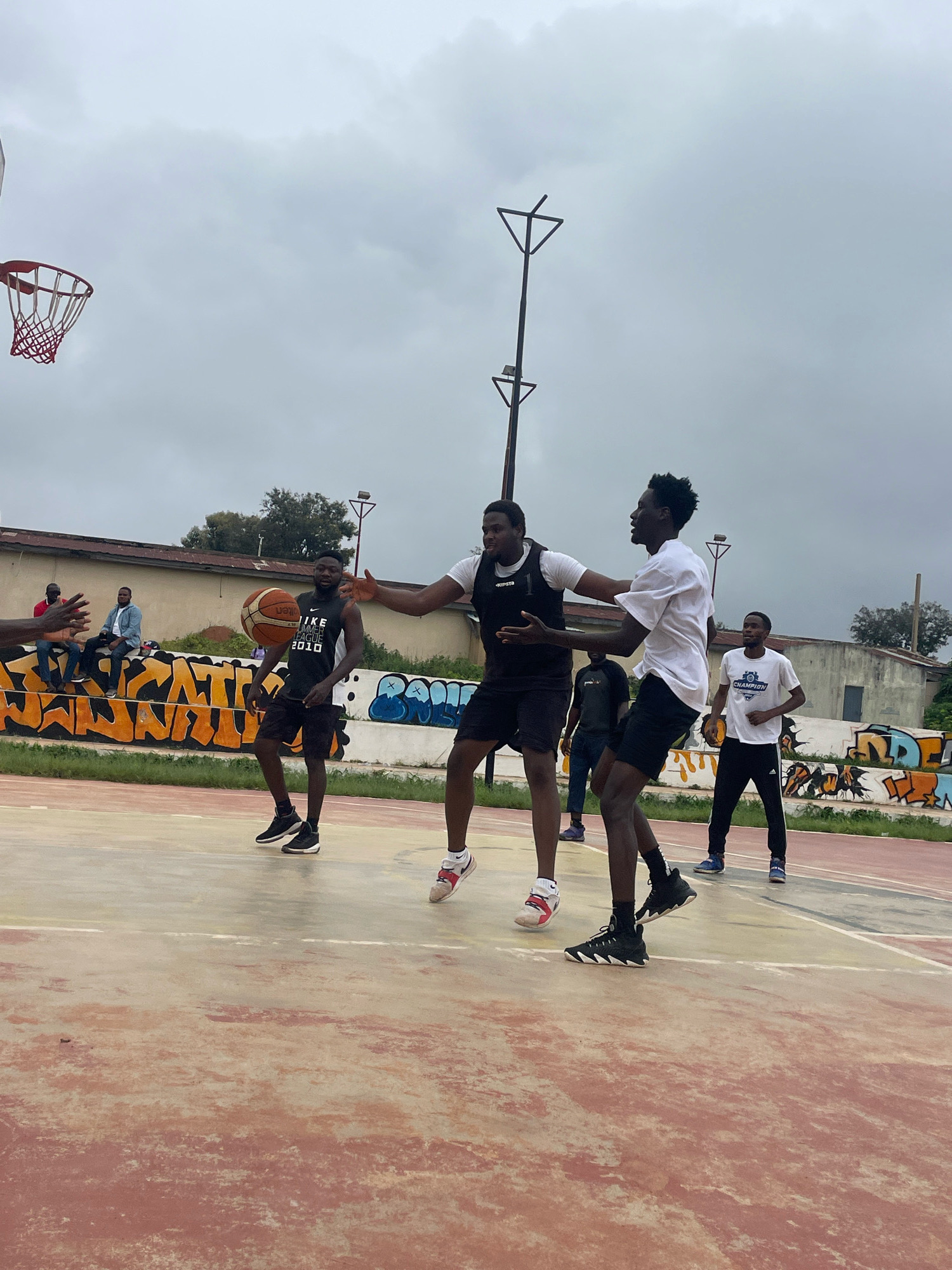 The image depicts an outdoor basketball court in the city of Gura Top, Nigeria, under an overcast sky. Four men are actively engaged in a basketball game on the worn, reddish-brown court. Two individuals are in the foreground, one wearing a white t-shirt and black shorts, the other a black tank top and black shorts, with one of them holding a basketball. Two other men are visible in the midground, one in a black tank top and black shorts dribbling the ball, and another in a white t-shirt and black pants standing on the court. In the background, several other people are seated on a low wall adorned with graffiti. The court is surrounded by buildings and some vegetation, and a basketball hoop with a red rim and white net is visible in the upper left corner.