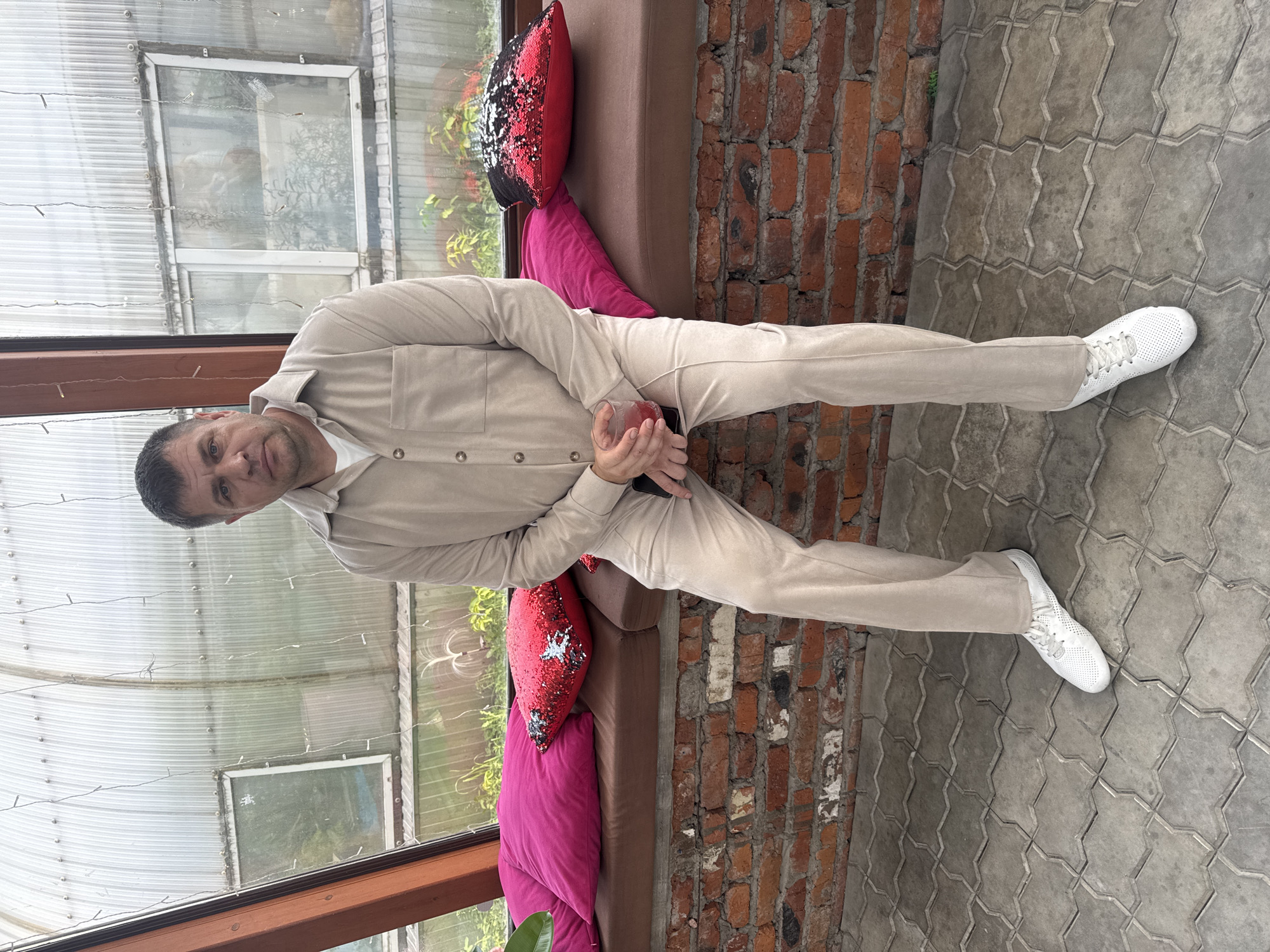 A middle-aged man with short, dark hair and a serious expression sits on a cushioned bench in what appears to be a greenhouse or conservatory. He is wearing a beige long-sleeved shirt and matching pants, with white sneakers. In his right hand, he holds a clear glass containing a pink liquid, and his left hand rests on his lap. The bench is upholstered in brown, and behind him are two large cushions: one bright pink and another red with black sequins. The background consists of a brick wall supporting the bench and a wall made of corrugated translucent material with windows, revealing greenery outside. The floor is paved with grey interlocking tiles. The overall impression is a casual, indoor setting. The specific location in Komsomolsk-on-Amur, Russia, cannot be determined from the image alone.