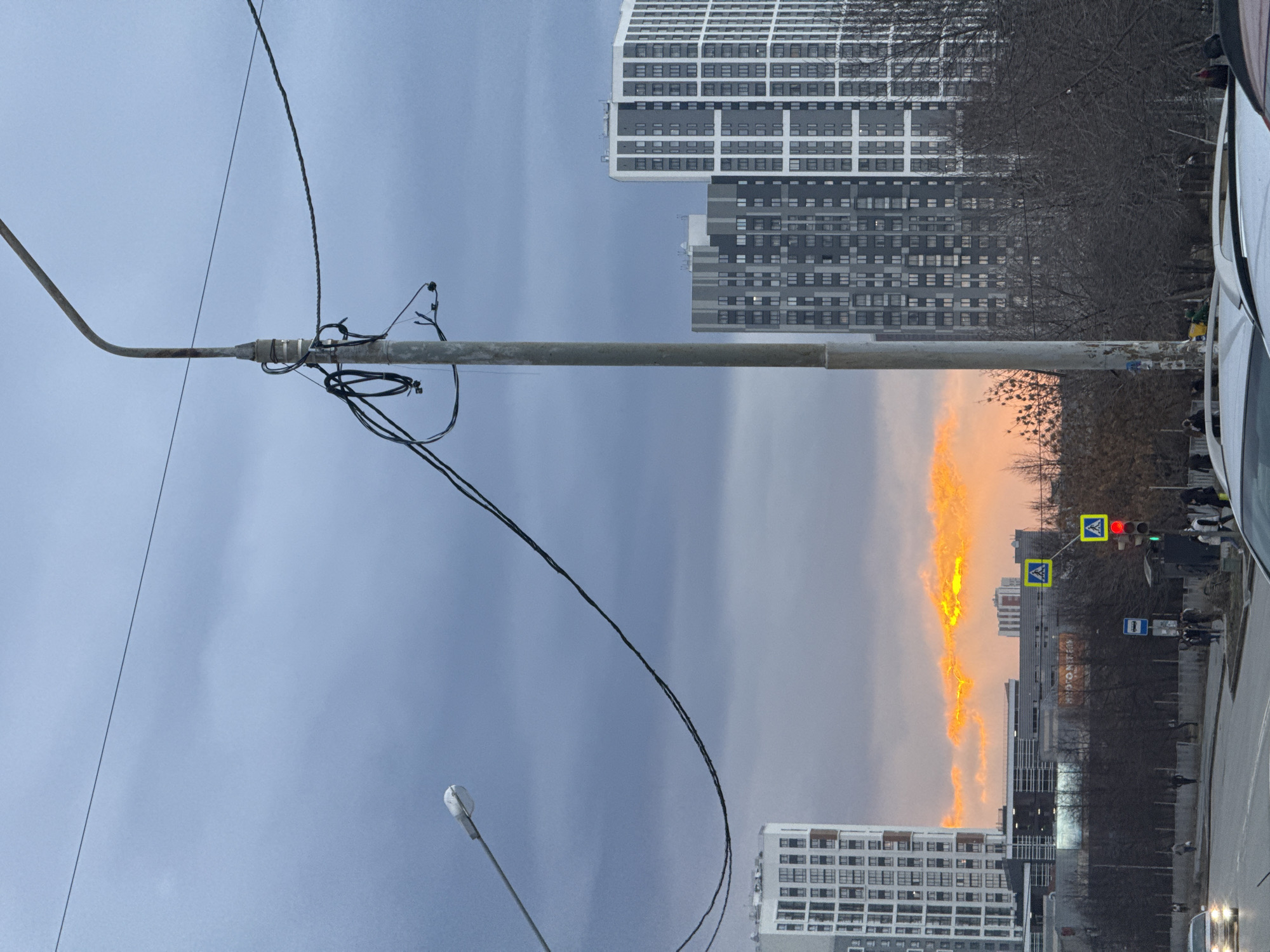 A daytime urban scene in Yekaterinburg, Russia. The viewpoint is at street level, looking up and slightly across the road. A utility pole is prominent on the left side of the frame, with multiple wires attached. In the background, there are several high-rise buildings with a modern architectural style. The sky is predominantly overcast with a mix of grey and blue tones. A bright orange light seems to emerge from behind the buildings, likely a sunset or a similar atmospheric effect. A pedestrian crossing sign, with a yellow and blue design, and a traffic light showing red, are also visible. People are present on the sidewalk. A portion of a car is visible on the right side of the frame, suggesting the photo was taken from a vehicle. The environment is generally urban, with a mix of built structures and natural elements such as bare trees.