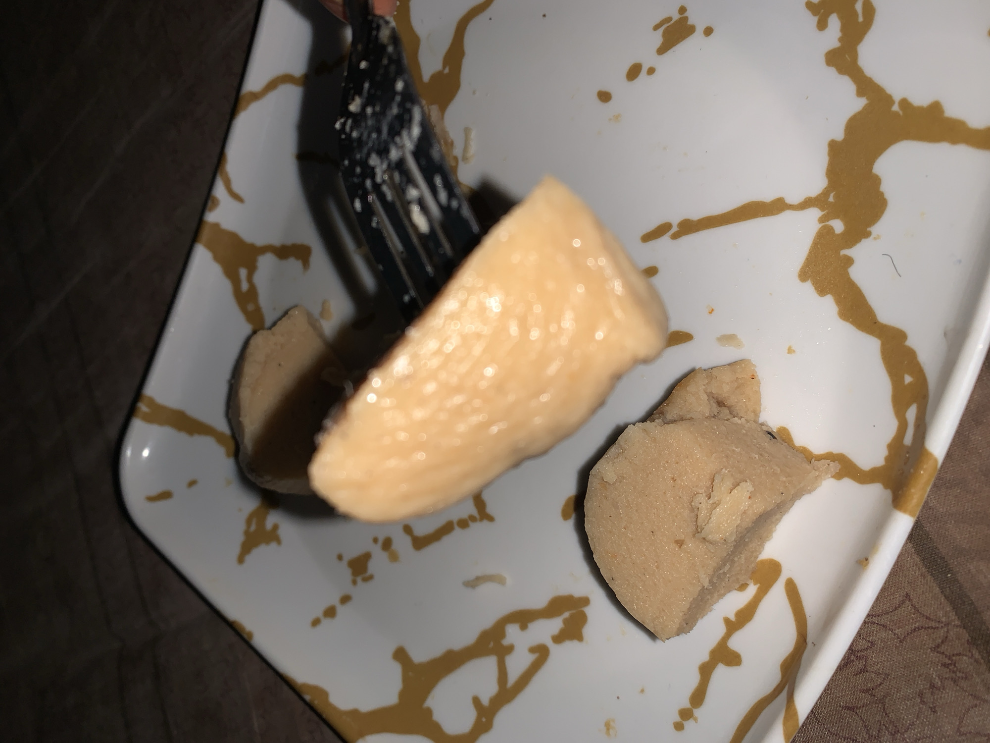 The image shows a white plate with a brown abstract design. On the plate are two pieces of a light brown, dough-like food. One piece is a large, elongated chunk, and the other is a smaller, wedge-shaped piece. A black fork is positioned on the plate, with some of the food stuck to its tines. The food appears to be some sort of paste or cooked dough, possibly fufu or a similar staple. The plate is set on a brown and white checkered surface, likely a tablecloth. The background is dark and out of focus, suggesting indoor lighting. The overall impression is of a meal being prepared or consumed. There is no specific indication of the location being Rafin Par, Nigeria.