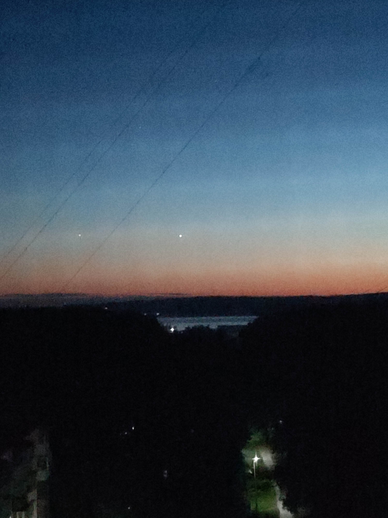 The image presents a serene twilight landscape, capturing the transition from day to night. The sky, dominating the upper half, displays a striking gradient, shifting from a deep indigo and dark blue at the top to warm orange and reddish hues closer to the horizon. Two distinct, bright white points of light, likely planets or bright stars, are prominently visible in the orange section of the sky. Several faint, diagonal lines, appearing to be power or utility lines, traverse the upper sky, indicating an urban or suburban vantage point, possibly from a building window or balcony.
Below the colorful sky, the horizon is marked by a dark, silhouetted landmass, likely dense trees or distant hills. A tranquil body of water, possibly a river or lake, stretches across the midground, reflecting the faint light and appearing as a slightly lighter band. A few scattered, distant lights can be seen along the far shore, suggesting distant settlements.
The foreground is enveloped in deep shadow, almost completely dark. On the far left, the subtle outline of a building is barely visible through the darkness. On the lower right, a single, brightly glowing streetlamp illuminates a small patch of ground below it, possibly a path or a grassy area. No people or specific activities are visible within the frame. There is no discernible text. The scene evokes a calm and peaceful atmosphere characteristic of dusk.