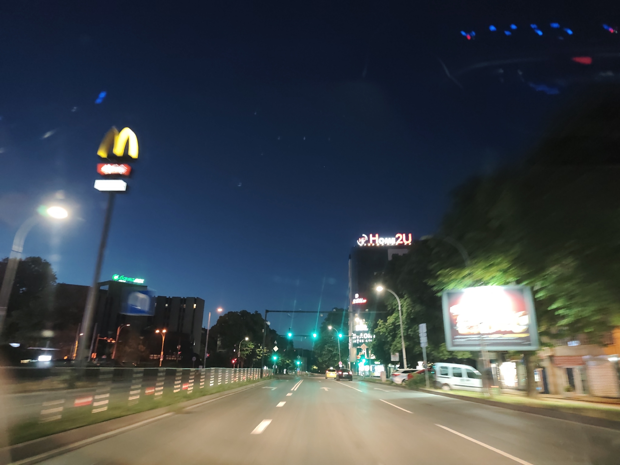 The image shows a view of a street in Varna, Bulgaria at night. The scene is captured from a moving vehicle. On the left side of the street, a McDonald's sign is prominently displayed. Across the street, a building with a "Home2U" sign is visible, along with a large billboard displaying a burger advertisement. Traffic lights are illuminated green, and several cars are on the road. Trees line the sides of the street, and streetlights provide illumination. The sky is dark blue, indicating it is nighttime.