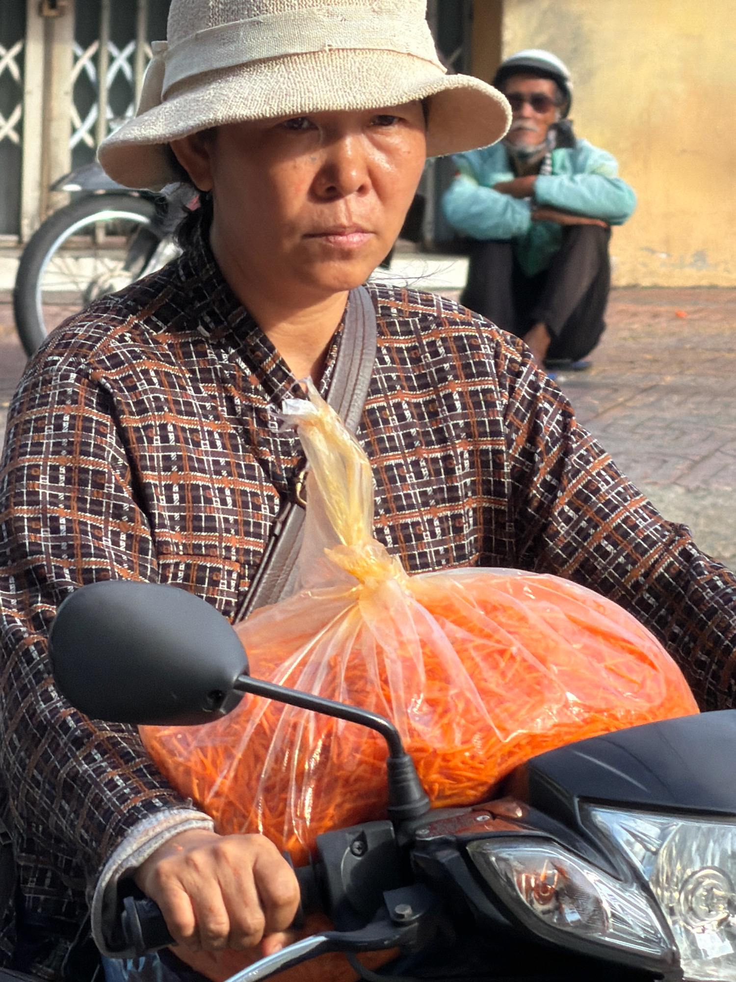 A woman with a light-colored straw hat is riding a motorcycle in what appears to be Nha Trang, Vietnam. She is wearing a brown and white plaid long-sleeved shirt and carrying a large clear plastic bag filled with shredded orange vegetables, likely carrots, in front of her. Her right hand is on the motorcycle's handlebar. In the background, a man wearing a blue shirt and a helmet is sitting on the ground. The scene is outdoors, with a building and a fence visible behind the people.