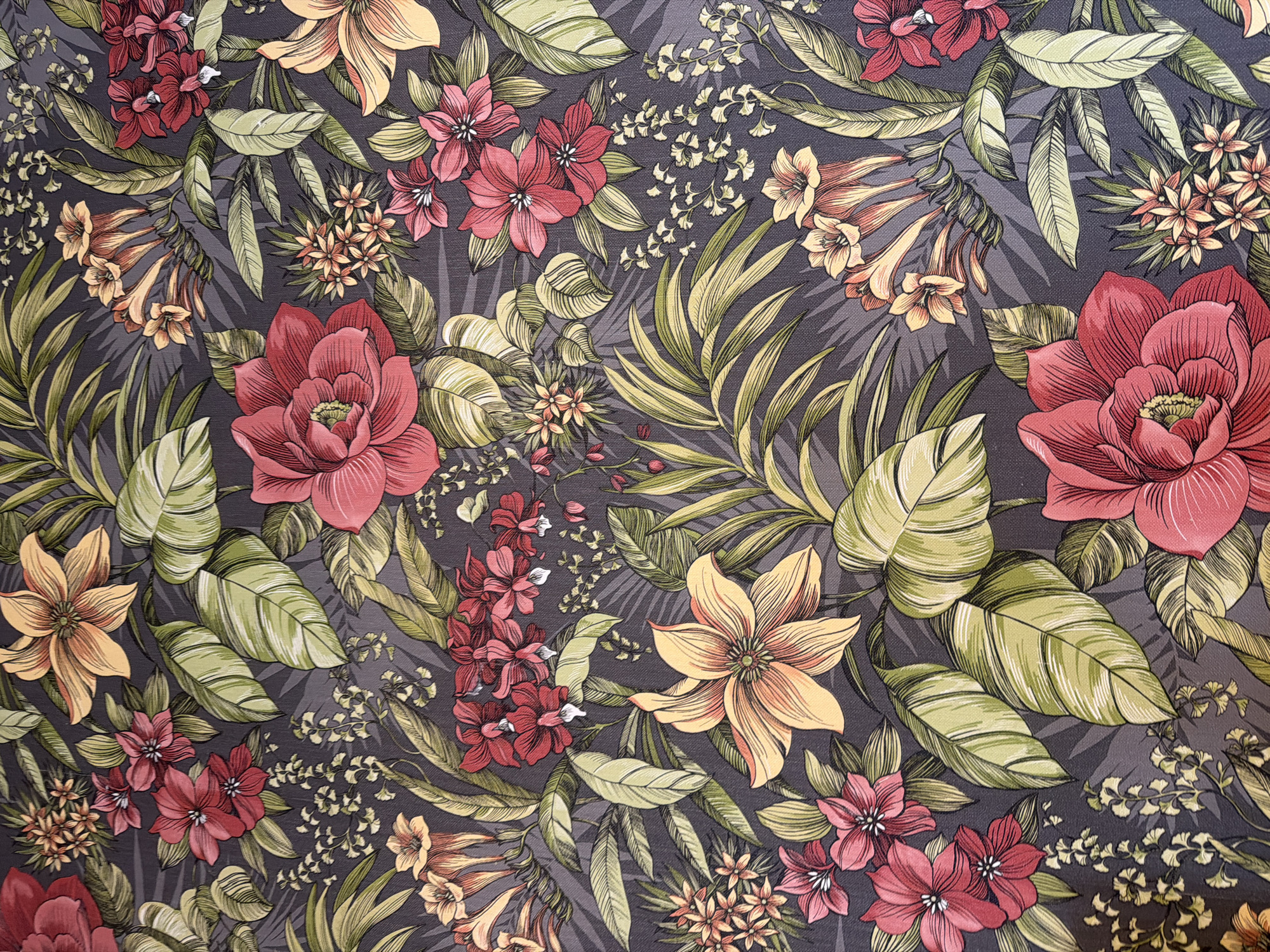This is a close-up, eye-level, full shot of a dark grey fabric with a vibrant, detailed floral and foliage pattern. The pattern features a variety of flowers in shades of red, pink, and yellow, interspersed with large, broad green leaves that have prominent lighter green veins. There are also smaller, delicate clusters of light yellow flowers and wispy, light green branches with tiny white blossoms. The overall impression is lush and tropical, with a high level of detail and shading contributing to a sense of depth and texture within the floral design. The background is a solid dark grey, which makes the colors of the flowers and leaves pop.