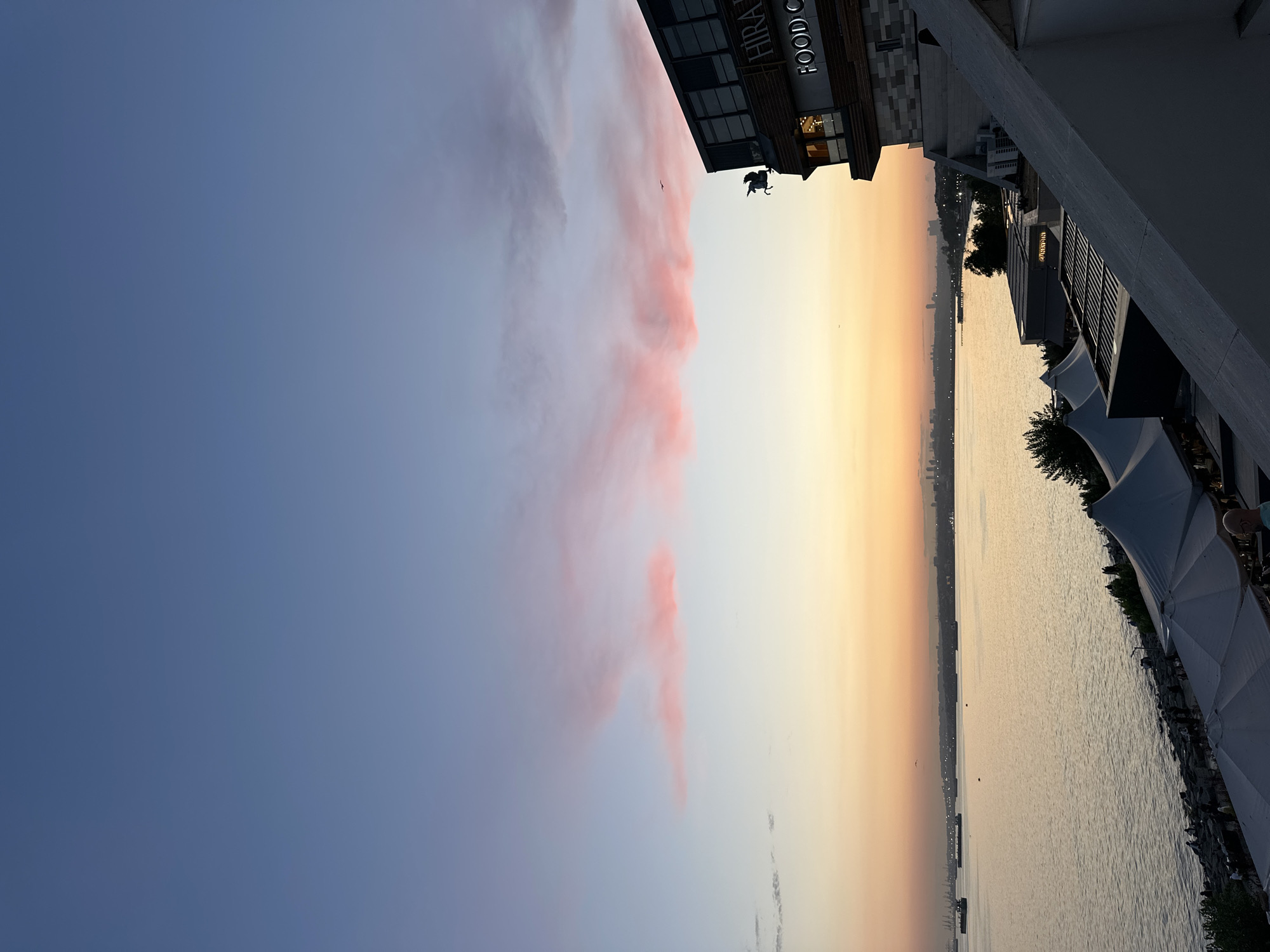 The image depicts a scenic view of the sea, likely the Marmara Sea, during sunset, with the skyline of Istanbul visible in the distance. In the foreground, a modern building with a sign reading "HIRA FOOD CO." is prominent, featuring dark-tinted windows and a unique architectural element of a silhouette of a person on horseback. Several white market tents are set up along the waterfront, suggesting a public space or a market area. Ships and cargo vessels are anchored in the sea. The sky is a gradient of blue and pink, characteristic of a sunset. The location is identified as being in Bakırköy, Turkey.