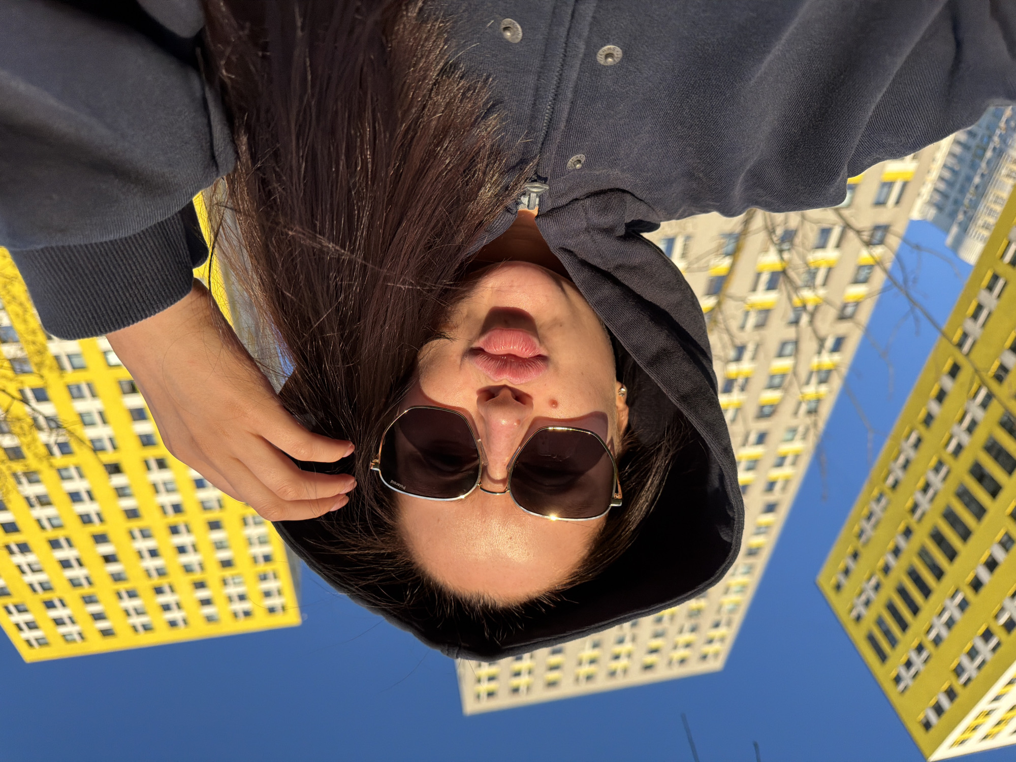 A woman wearing a hooded navy zip-up sweatshirt and sunglasses looks down at the camera. Her hair is dark and straight. Her left hand is near her hair. Behind her are yellow and white high-rise buildings against a blue sky. The scene is located in the city of Khimki, Russia.