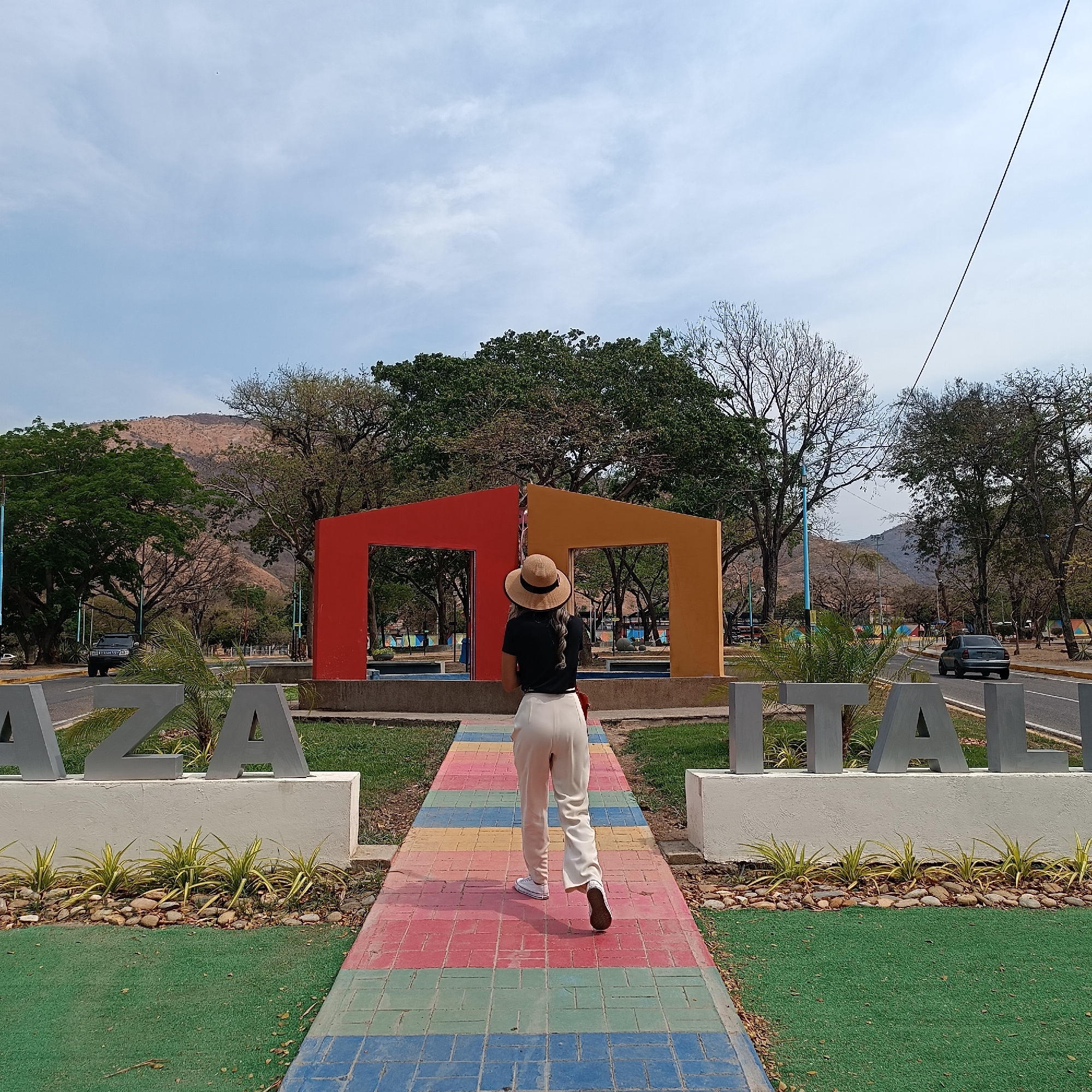 A photograph depicts an outdoor scene within Plaza Italia in La Victoria, Venezuela, on what appears to be a clear day with a partly cloudy sky. The perspective is from behind a single individual walking away from the camera, directly towards the center of the plaza.
The subject is a person wearing a light brown straw hat with a dark band, a black short-sleeved top, light-colored (likely white or cream) trousers, and white sneakers with brown soles. A small red object is visible on their right hip. Their long, wavy, light-colored hair is partially visible beneath the hat. They are positioned centrally on a multicolored paved pathway composed of rectangular blocks in alternating horizontal stripes of red, blue, green, and yellow.
In the mid-ground, at the end of the pathway, stands a prominent two-part sculptural structure. The left section is a red-orange, open-frame house-like shape, joined in the center to a right section of golden-orange, similarly open-frame structure. This structure is situated on a low, dark-colored base.
Flanking the pathway in the foreground are patches of green artificial turf, with planters containing small, spiky yellow-green plants and river stones. Large, grey, three-dimensional letters spelling "PLAZA" are visible to the left of the pathway, and "ITALIA" to the right, mounted on low, white pedestals.
Beyond the central sculpture, the plaza extends with additional green spaces and numerous trees. Some trees possess full green foliage, while others exhibit bare branches. Two paved roadways run parallel to the central plaza area, separated by grassy verges. On the left road, a dark-colored SUV (resembling a Hummer H3) is observed moving away from the camera. On the right road, a light blue or grey sedan is also moving away. Blue-green lampposts are scattered along the roadsides.
In the far background, arid, light brown mountains rise under the sky. A thin, black power line or cable stretches diagonally across the upper right quadrant of the image. A very small, dark speck is faintly visible in the upper central sky.
