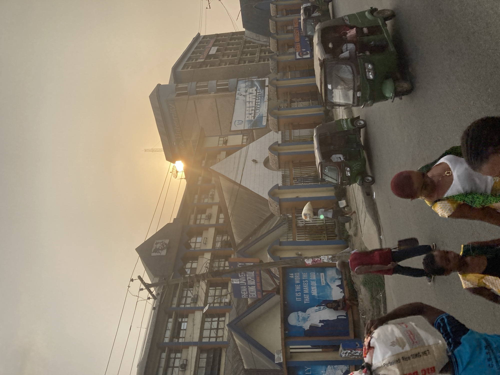 The image captures a street scene in Aba, Nigeria, prominently featuring Rhema University Teaching Hospital buildings. The buildings are multiple stories high, constructed in a combination of concrete and brick with blue and yellow architectural accents. In the foreground, pedestrians are walking along the street, and multiple green auto-rickshaws, known as "keke napep" in Nigeria, are driving. An advertising billboard for Rhema University is visible on the side of the building. The sky is overcast and the sun is setting, casting a haze over the scene.