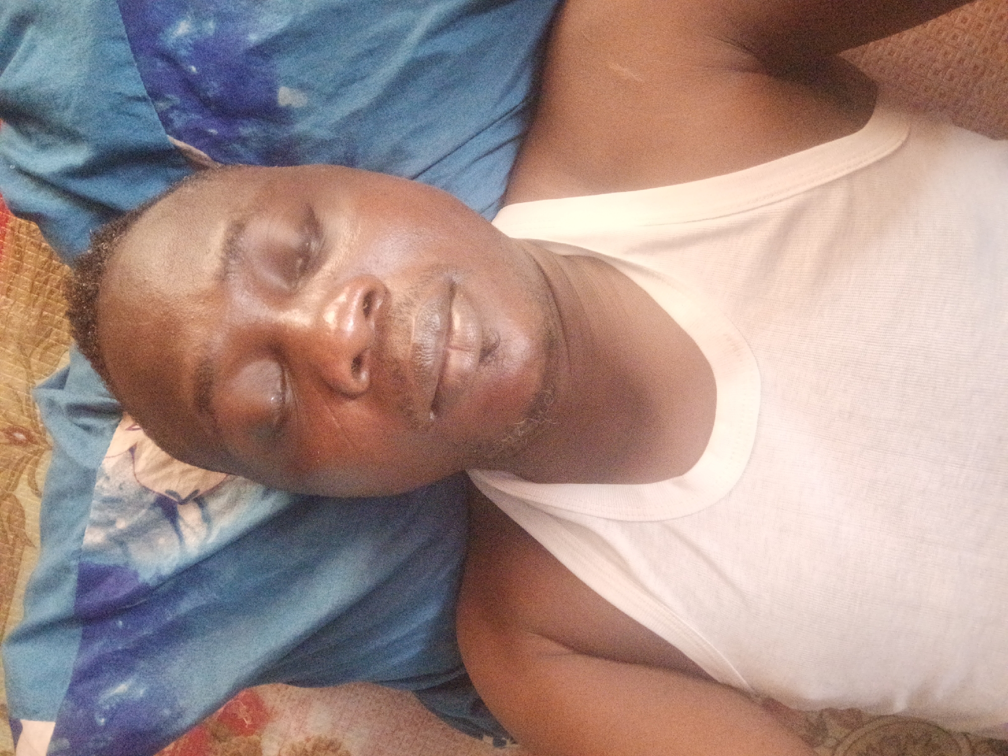 A man is lying on a bed in an indoor setting. He is wearing a white tank top. His eyes are closed and he appears to be sleeping. The bed has a blue and brown patterned blanket. The location is unknown.