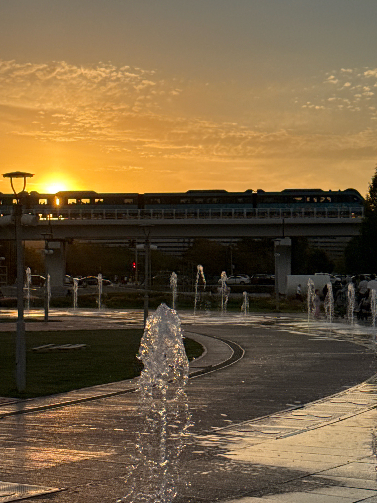 The image depicts a sunset scene at the Bayterek monument area in Astana, Kazakhstan. In the background, a train is visible on an elevated track, illuminated by the golden light of the setting sun. The sky is filled with scattered clouds, painted in hues of orange and yellow. In the foreground, a pathway with water fountains creates a dynamic element, with water jets spraying upwards, catching the light. The ground appears wet, reflecting the warm sunset colors. Several people are faintly visible in the background, near the fountains and along the pathway, suggesting a public gathering or recreational activity. The overall atmosphere is serene and picturesque, with the architectural elements of the city integrated with natural light and water features.