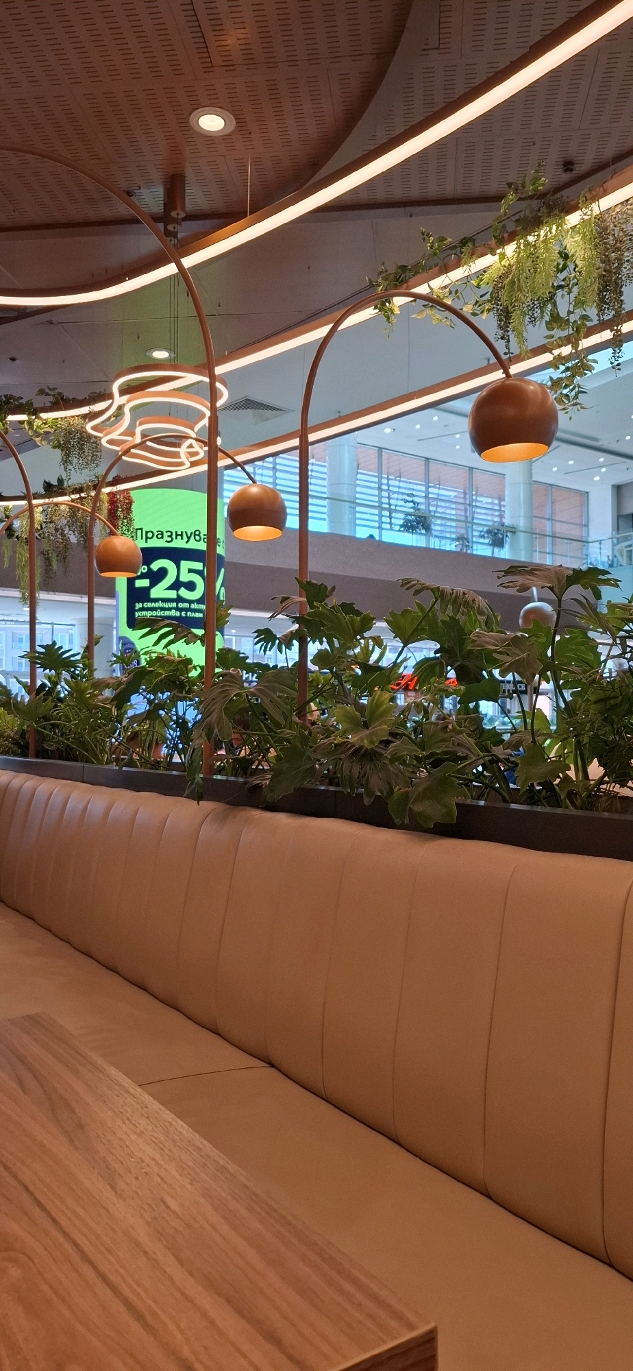 A vertical, medium shot captures an interior scene with a focus on a light brown upholstered booth and a wooden table in the foreground. Beyond the booth, a lush green plant display is visible, with large, leafy plants. Behind the plants, a bright green digital screen displays text and a "-25%" graphic, suggesting a sale or promotion. The background reveals a brightly lit, multi-level interior space, likely a shopping mall, with glass railings and architectural elements. Several decorative pendant lights, with copper-colored spherical shades, hang from arched structures above the plants. Green artificial vines are draped around the arches and along the ceiling. The overall atmosphere suggests a modern, well-lit commercial or dining space, possibly in Sofia, Bulgaria, given the context provided. No people are clearly visible in the frame.