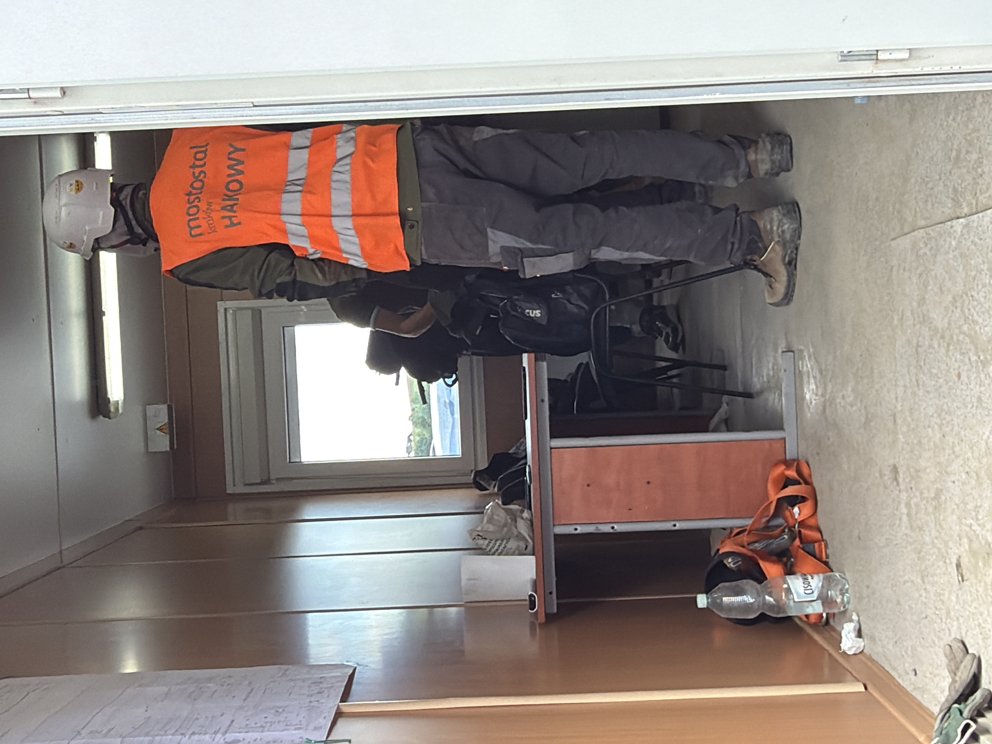 The image depicts the interior of a portable building, likely a temporary site office or changing room, at a construction location in Stróża, Poland. The scene is occupied by two individuals, both wearing work attire.
In the foreground, a person is viewed from the back, wearing a white hard hat, an orange high-visibility vest with reflective stripes, and dark work trousers. The vest has text printed on the back, including "mostostal kraków" and "HAKOWY". This person is also wearing sturdy brown work boots.
Behind this individual, another person is partially visible, also in work clothes. They are standing near a window and a desk.
The portable building has wooden paneling on the walls and a simple metal-framed desk with an orange front panel. On the desk, there are various items, including a white paper roll, a crumpled plastic bag, and what appears to be a small electronic device. On the floor next to the desk is a large plastic bottle of water and an orange safety harness. A second backpack is visible near the person in the background.
The overall environment appears functional and utilitarian, consistent with a construction site. The lighting suggests it is daytime, with natural light entering through the window and artificial light from a fixture visible on the ceiling. The floor is concrete.