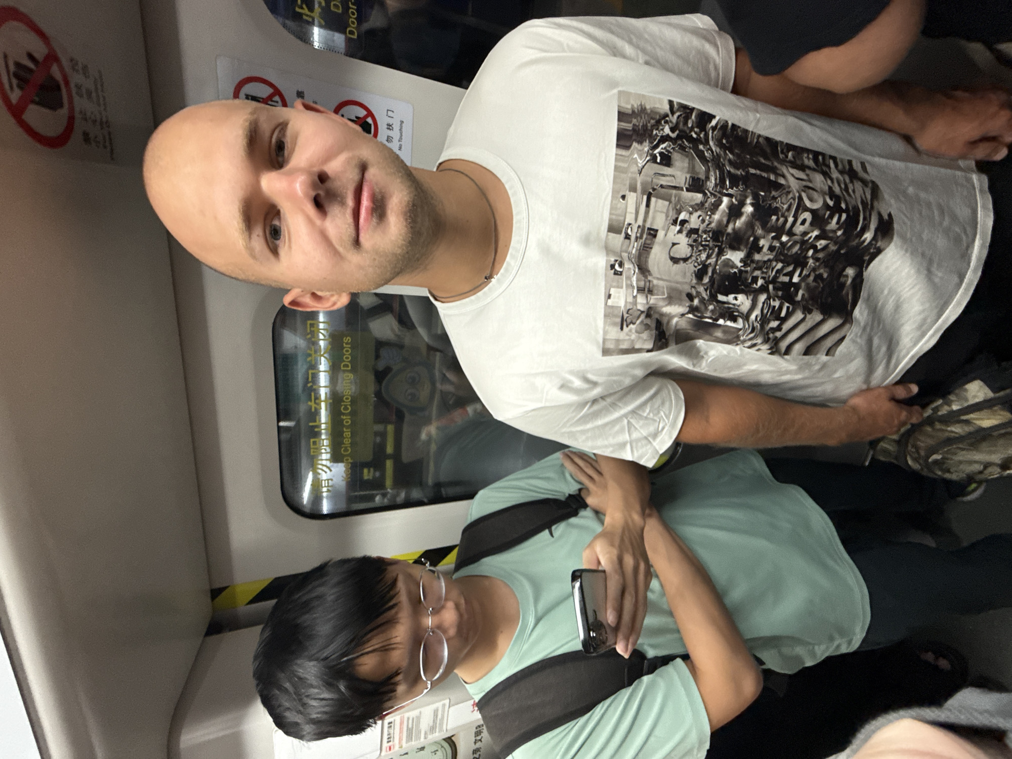 Two young adult males are standing inside a subway train car, likely in Tianhe District, China, given the visible Chinese text on signs. The male on the left, with black hair and glasses, is wearing a light green t-shirt and a black backpack. He is looking down at his smartphone, which he holds in his left hand. His arms are crossed. The male on the right, who is bald and has fair skin, is wearing a white t-shirt with a graphic design and a necklace. He is looking towards the camera with a neutral expression. The subway car interior is visible, with seats and handrails. Signs with Chinese text and pictograms are affixed to the walls and doors, one of which says "Keep Clear of Closing Doors" in English. The lighting is artificial, typical of an indoor transit environment.