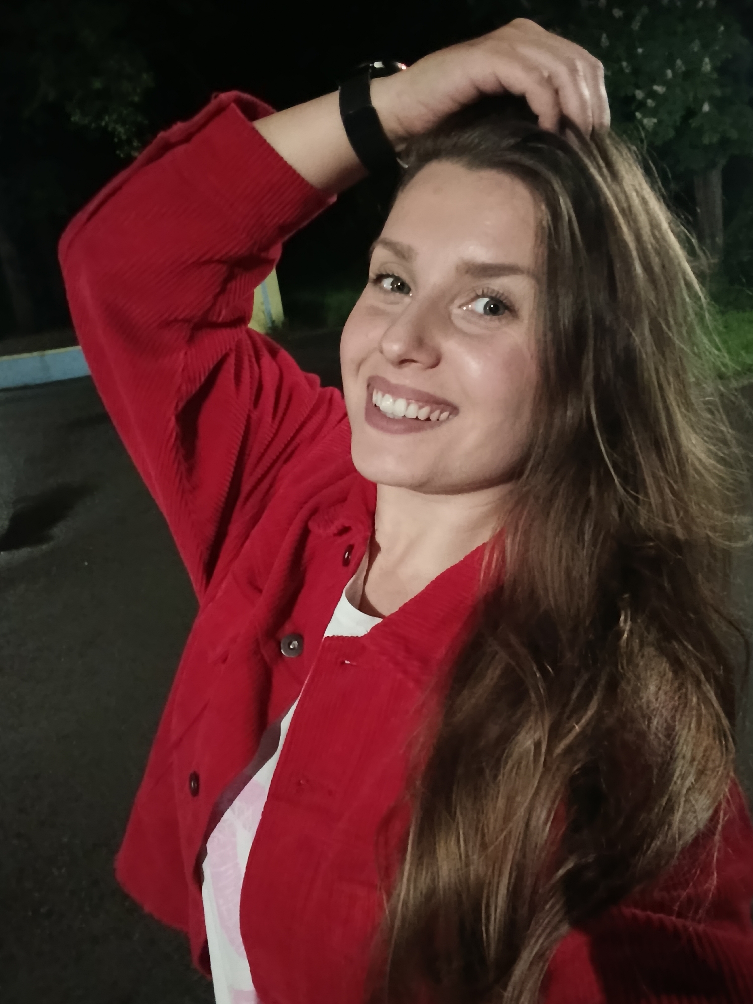 A young woman stands outdoors, smiling and looking towards the camera. She has long brown hair and is wearing a red corduroy jacket over a white t-shirt. Her right arm is raised, with her hand touching her hair. A smartwatch is visible on her left wrist. The background is dark, suggesting it is nighttime, and includes trees and a building or structure. The location is within the city of Targovishte, Bulgaria.