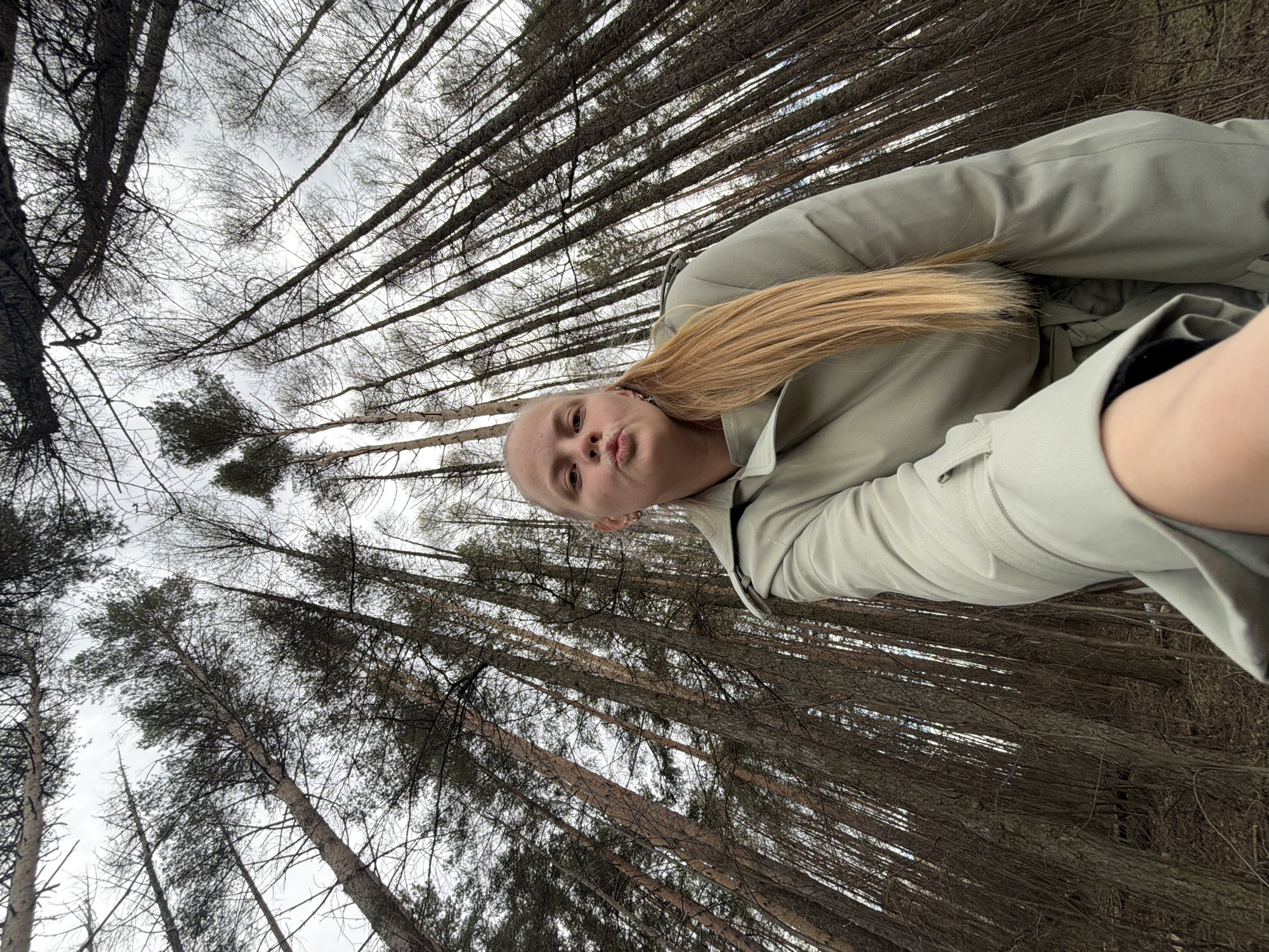 A selfie-style photo shows a person with light skin and blonde hair, wearing a light-colored coat or jacket. The person has their lips pursed in a playful manner. The background consists of a dense forest of tall, thin trees, with the sky visible through the branches. The trees appear to be without leaves, suggesting it might be winter or early spring. The exact location within Izhevsk, Russia, is difficult to ascertain based solely on this image.