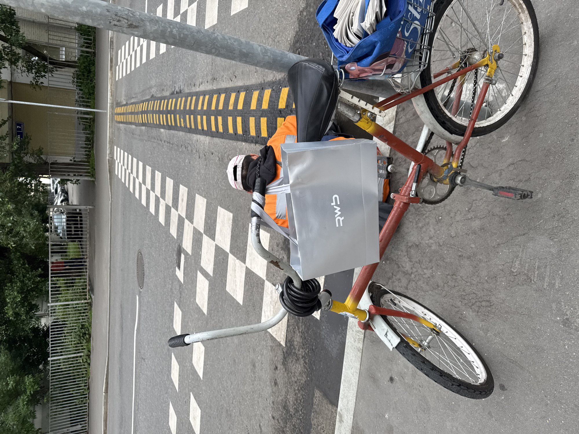 The image captures a close-up view of a red and yellow bicycle parked on an asphalt road in Moscow, Russia, near a crosswalk and a speed bump. The bicycle is secured with a black U-lock wrapped around the handlebars. A silver shopping bag with the logo "rws" is hanging from the handlebars. Behind the bicycle, a person wearing an orange high-visibility vest and a pink baseball cap is seated on the ground, facing away from the camera. The background shows a metal fence, trees, and buildings. The overall scene appears to be an outdoor urban environment.