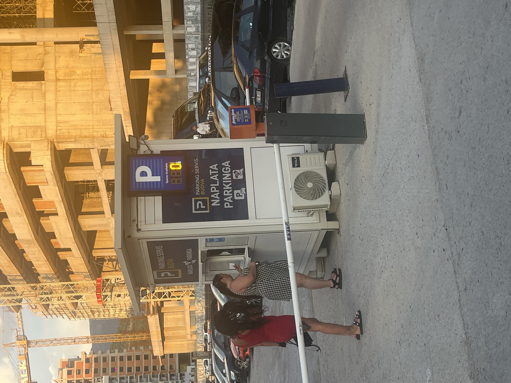This image shows a parking payment booth with two women standing near it. The woman on the left is wearing a black and white checkered dress and black sandals. The woman on the right is wearing a red dress and black sandals. Both women appear to be in conversation.
The parking booth has signs with "PARKING SERVIS BUDVA NAPLATA PARKINGA" and a "P" symbol indicating parking. One sign also displays "Spaces Available 880". An air conditioning unit is mounted on the side of the booth.
In the background, there is a construction site with concrete structures and cranes. Several cars are parked behind the booth, including a black car with a license plate that is partially visible. The scene appears to be outdoors, likely during the daytime, as indicated by the strong sunlight casting shadows. The lighting suggests it might be late afternoon or early morning. The overall setting is a city or a busy area with a mix of modern construction and existing infrastructure. The emotions of the people are not clearly discernible, but they appear to be engaged in a casual interaction.