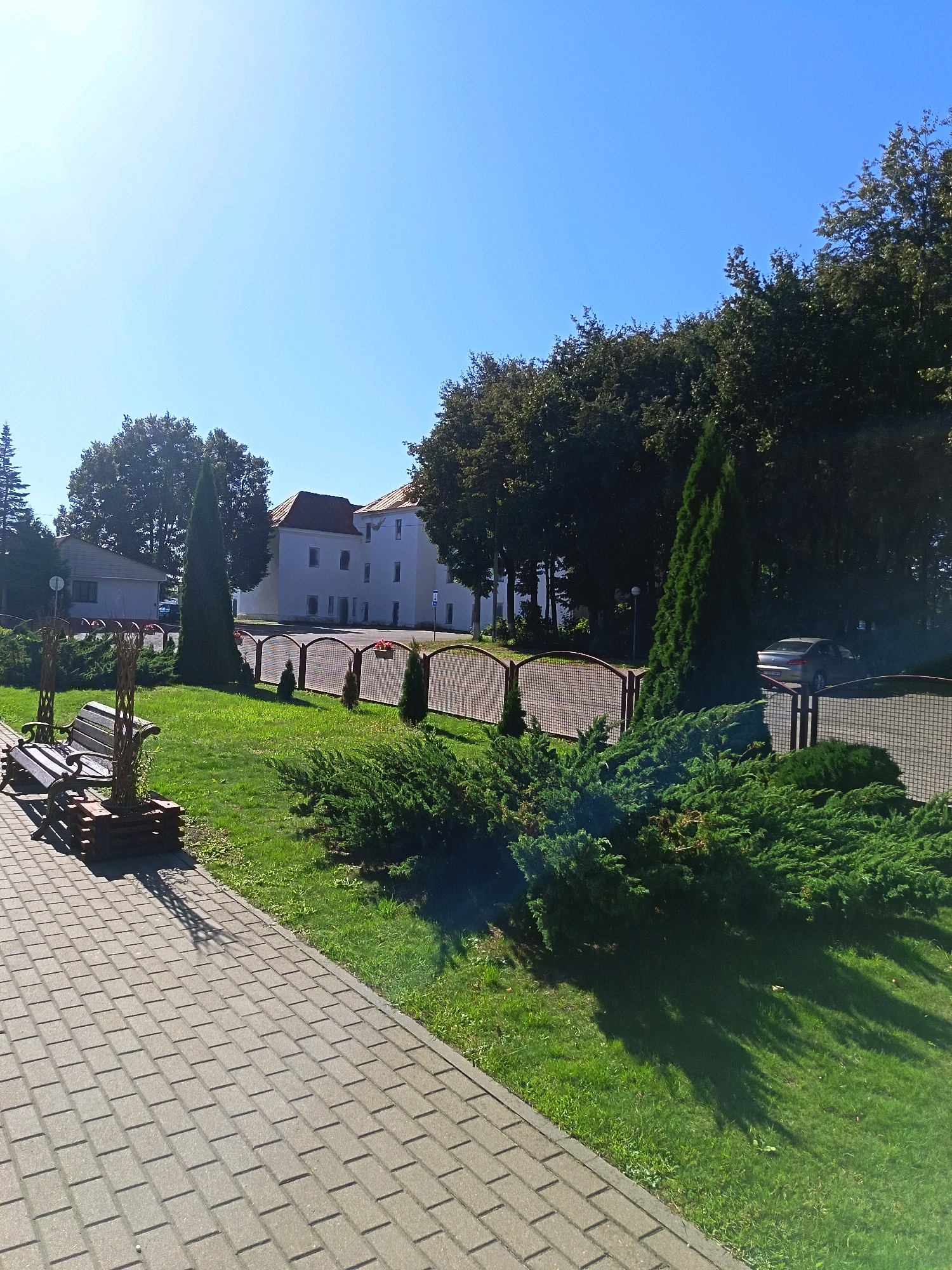 The image captures a sunny day scene with a clear blue sky. In the foreground, a paved pathway made of grey rectangular bricks leads diagonally across the frame. To the left of the pathway, a wooden bench is situated on a patch of green grass, with a small decorative wooden structure next to it. A low, manicured hedge runs along the edge of the grass.
Behind the hedge, a fence with arched brown metal posts and wire mesh separates the grassy area from a larger paved space. Beyond the fence, several evergreen trees, including a tall, slender cypress, are visible. In the background, a multi-story white building with a red-tiled roof stands under a bright sky. A silver car is parked in front of the building, partially obscured by trees and a fence. The overall environment appears to be an outdoor public space, possibly a park or the grounds of an institution in Niasvizh, Belarus.