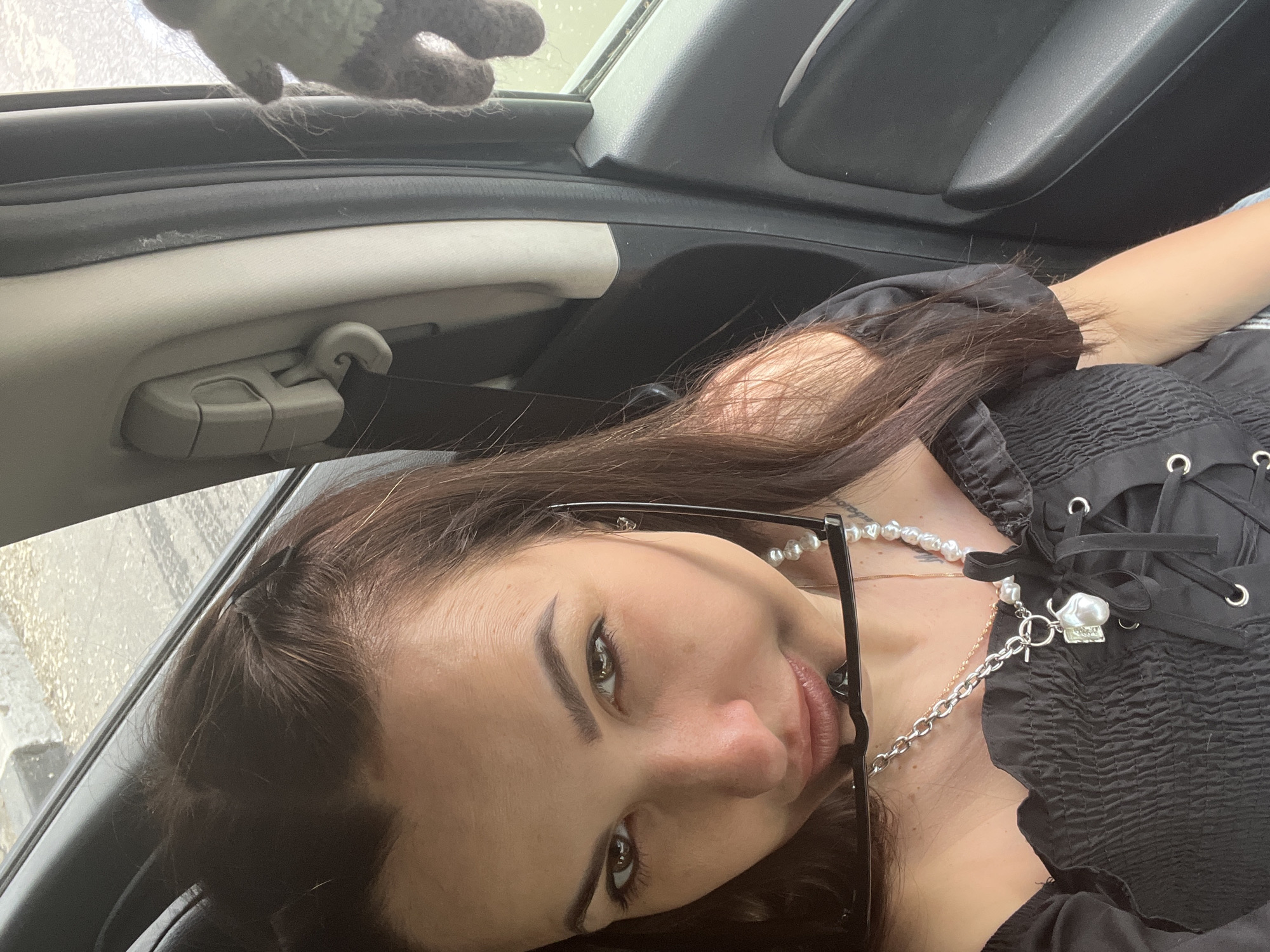 A young woman is captured in a tilted, close-up shot, appearing to lie down inside a car. Her long, dark brown hair spreads out around her head and shoulders. She looks directly at the camera with a calm expression and a faint smile. Her eyes are light-colored, possibly green or hazel, and are emphasized with dark eye makeup, complemented by prominent dark eyebrows. Black-framed eyeglasses rest on her face, seemingly pushed down.
She is dressed in a black top featuring shirred or smocked fabric on the bodice and full sleeves. The front of her top has a lace-up detail secured with silver eyelets. Around her neck, she wears a necklace made of irregularly shaped pearls, from which a silver chain extends, adorned with a toggle clasp and a distinctive baroque pearl or shell-like charm.
The background reveals the interior of a car, including a light-colored ceiling or pillar trim, a grey clip or hook, and various dark grey or black car panels. A blurred view through a car window indicates an outdoor setting, possibly under overcast conditions. In the upper left portion of the image, a fuzzy, light-colored object with dark patches, resembling a stuffed animal or a textured glove, is partially visible. The scene is illuminated by natural daylight. No text is visible in the image.